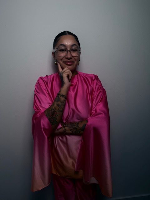 Woman in pink dress with glasses and tattoos, standing against a plain background, smiling and touching her face.