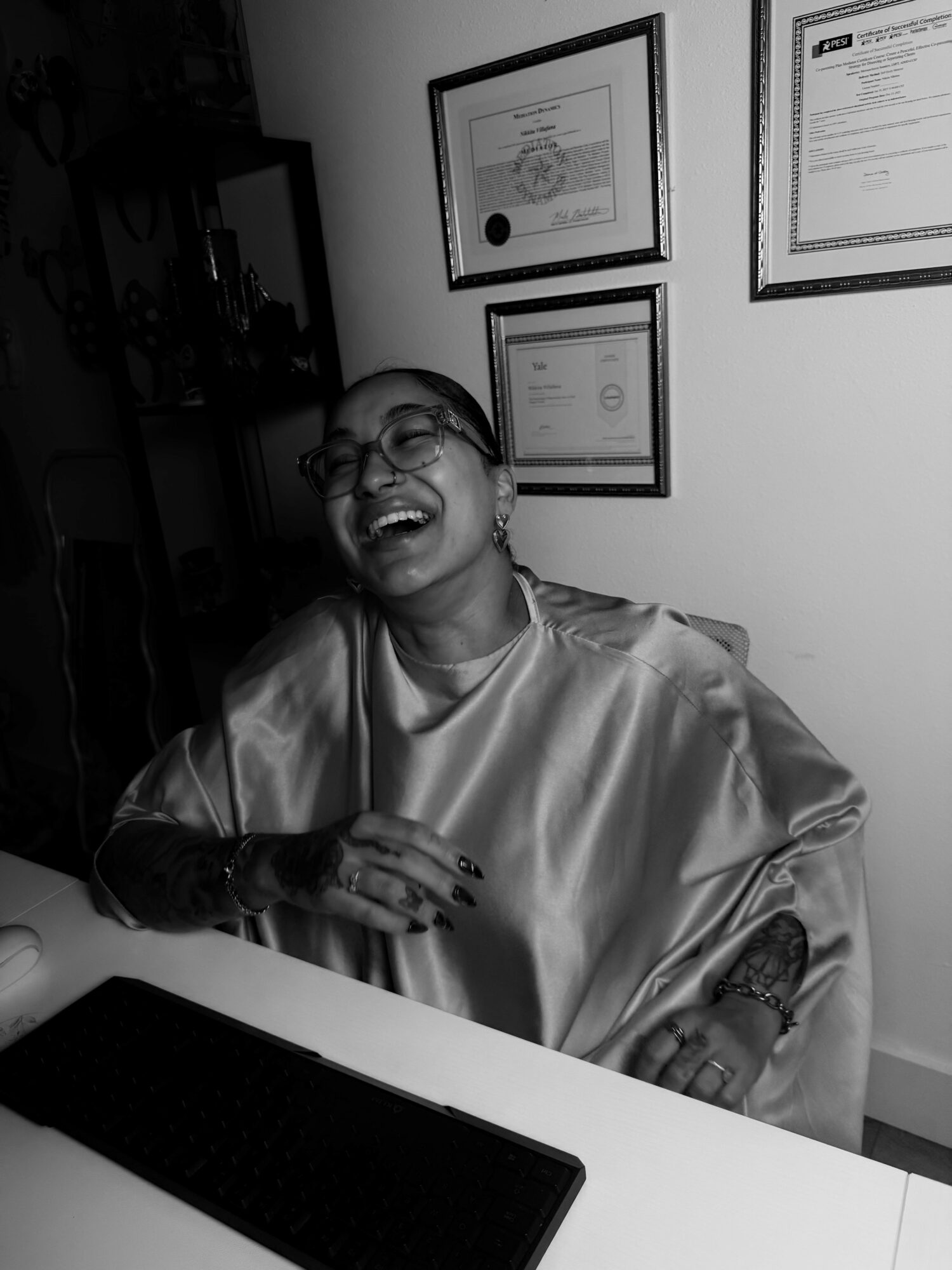 Person taking a selfie in a mirror, holding a phone, smiling, wearing a shirt, with framed certificates on the wall.