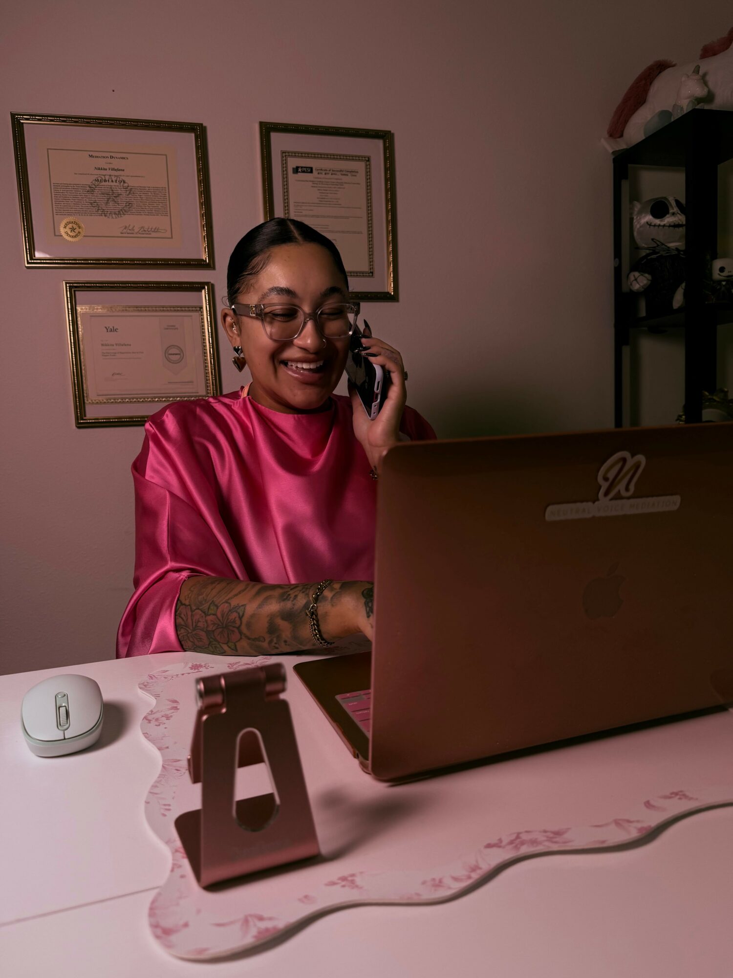 Woman in pink scrubs talking on phone at desk with certificates on wall, pink desk mat, and computer mouse.