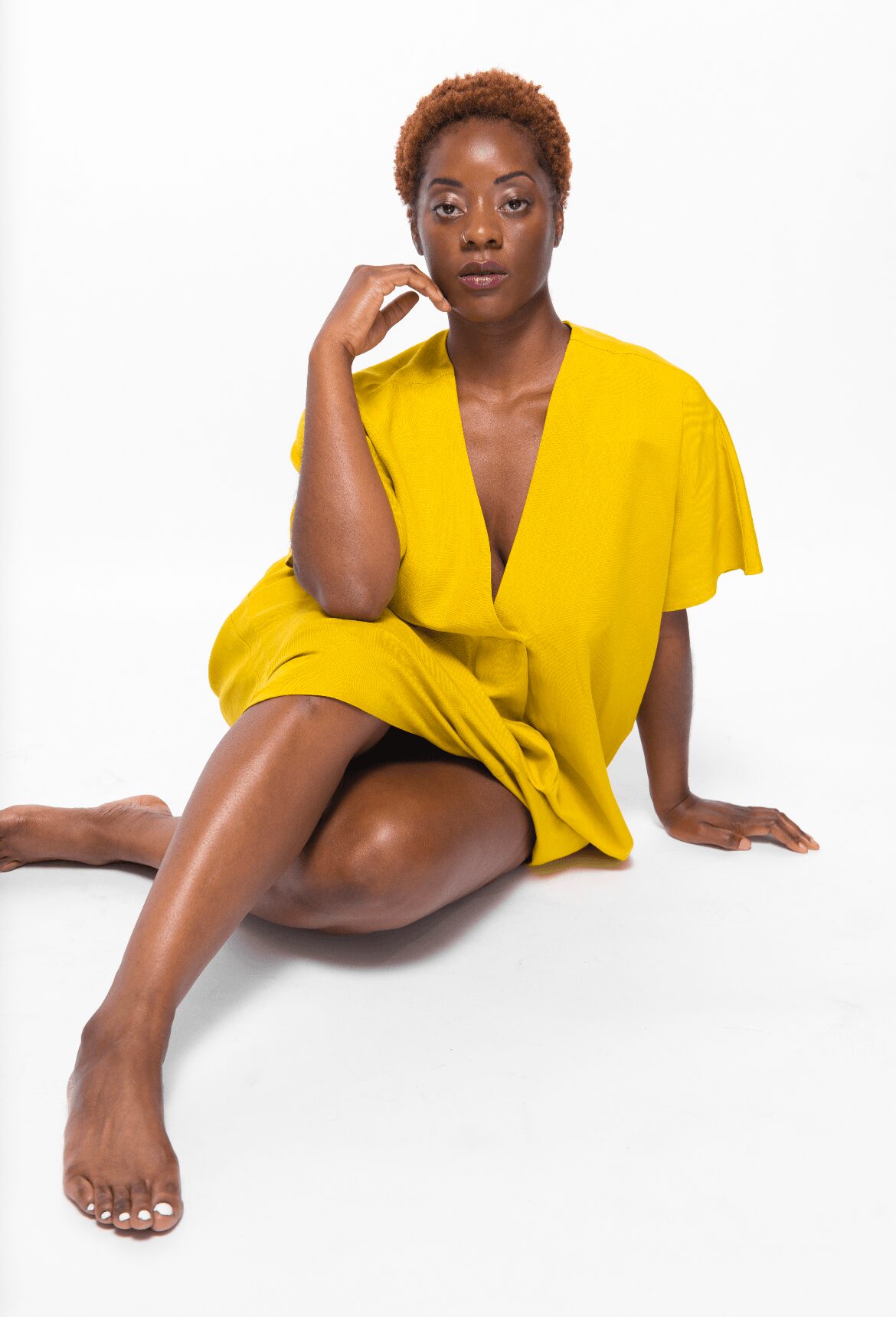 Woman in yellow dress sitting on the floor with one leg extended and hand resting on her face, white background.