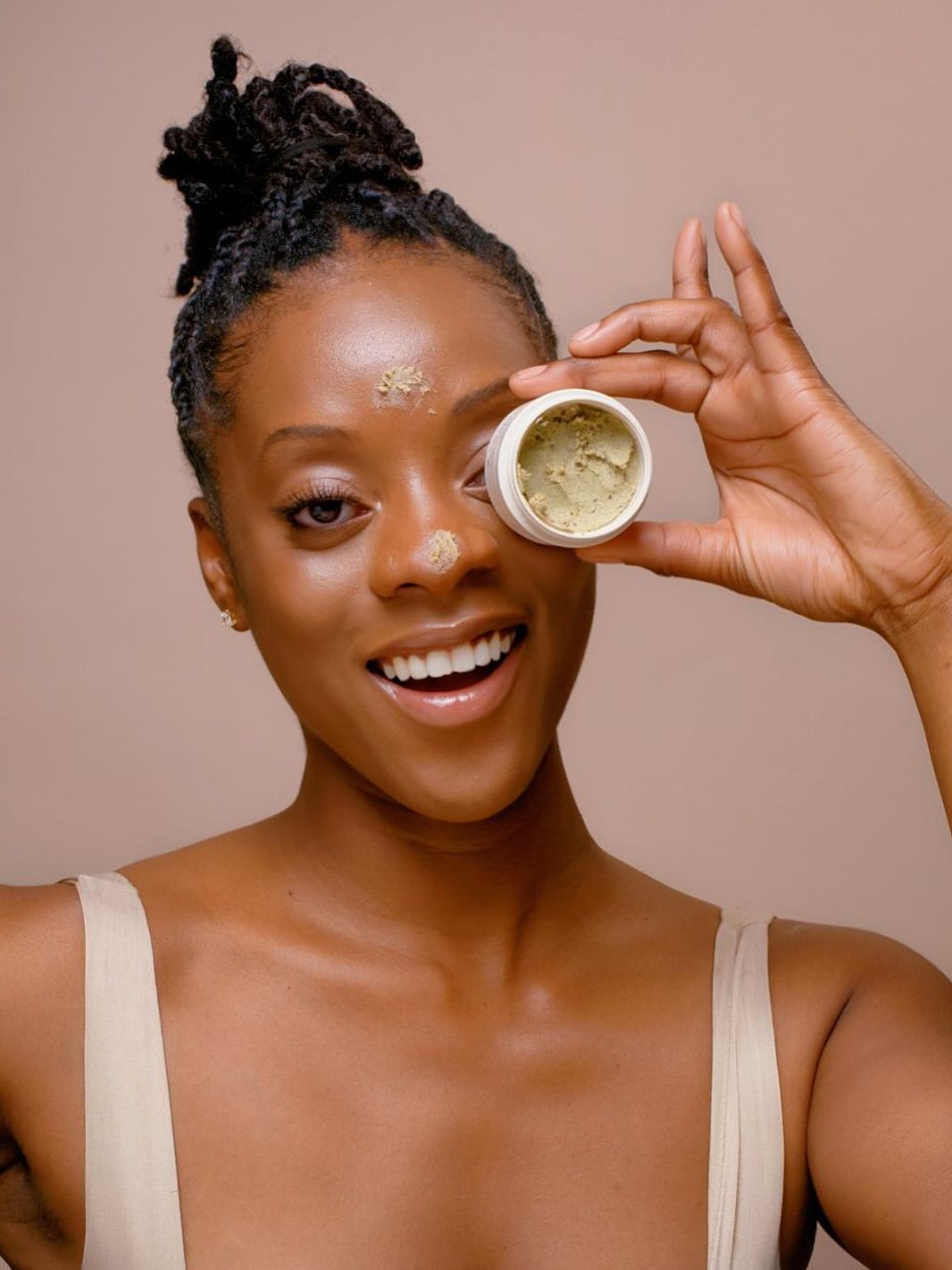 Young woman with natural hair holds a small container near her eye, smiling, against a neutral background.