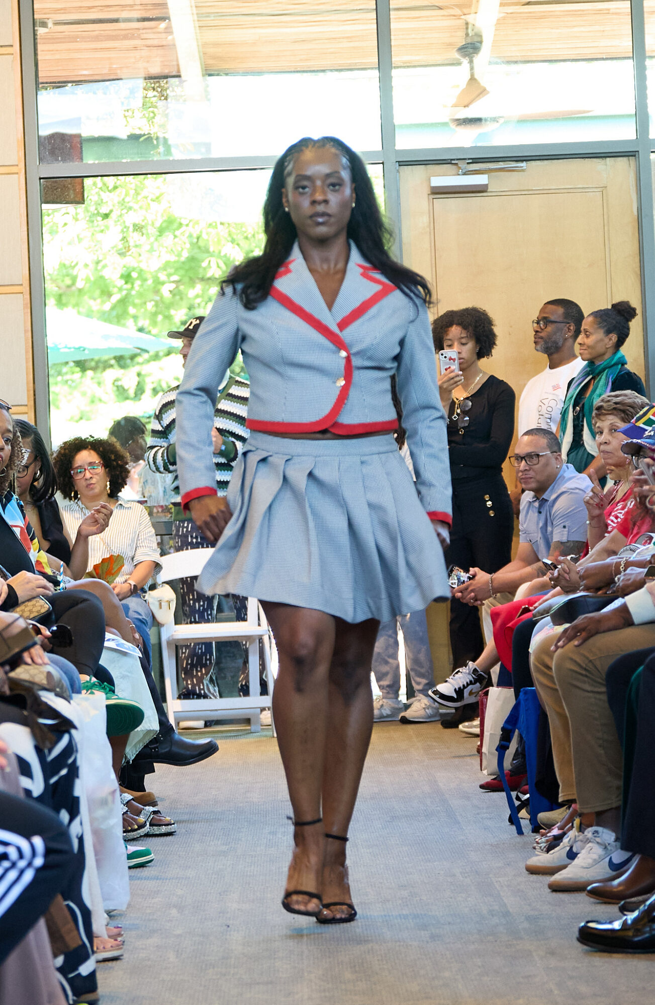 Female model walking down runway in a light blue pleated skirt and matching blazer with red accents, audience watching.