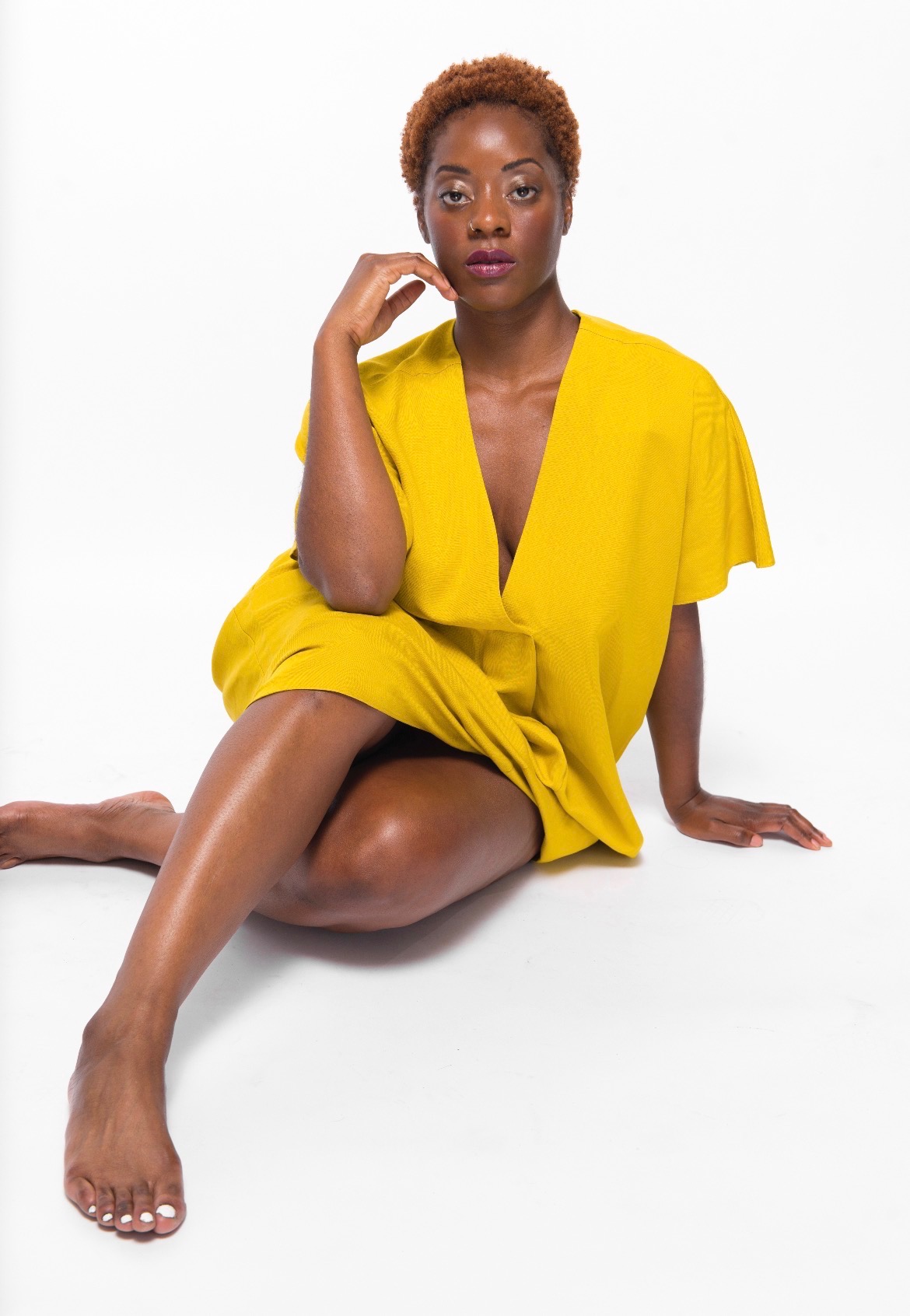 Woman in yellow dress sitting on the floor with one leg extended and hand near face, white background.