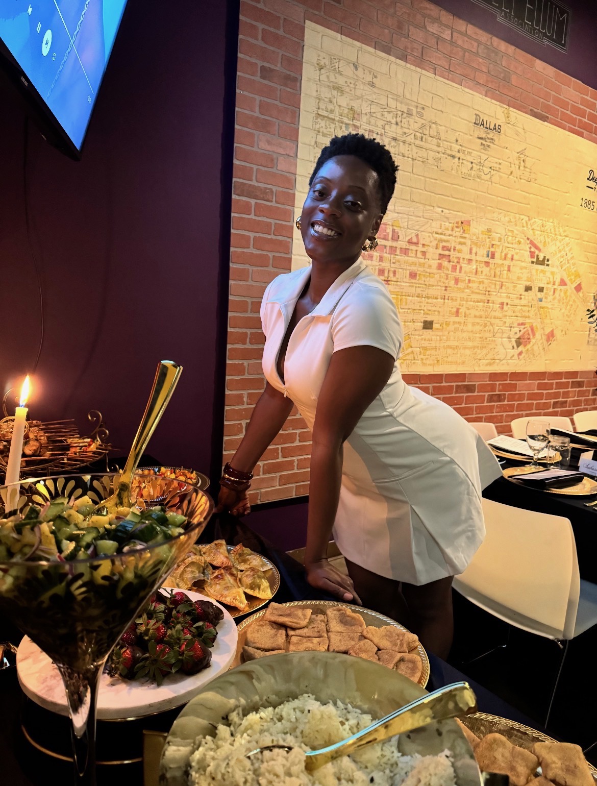 Smiling woman in white dress leaning over a table with food and a lit candle, in a restaurant setting.