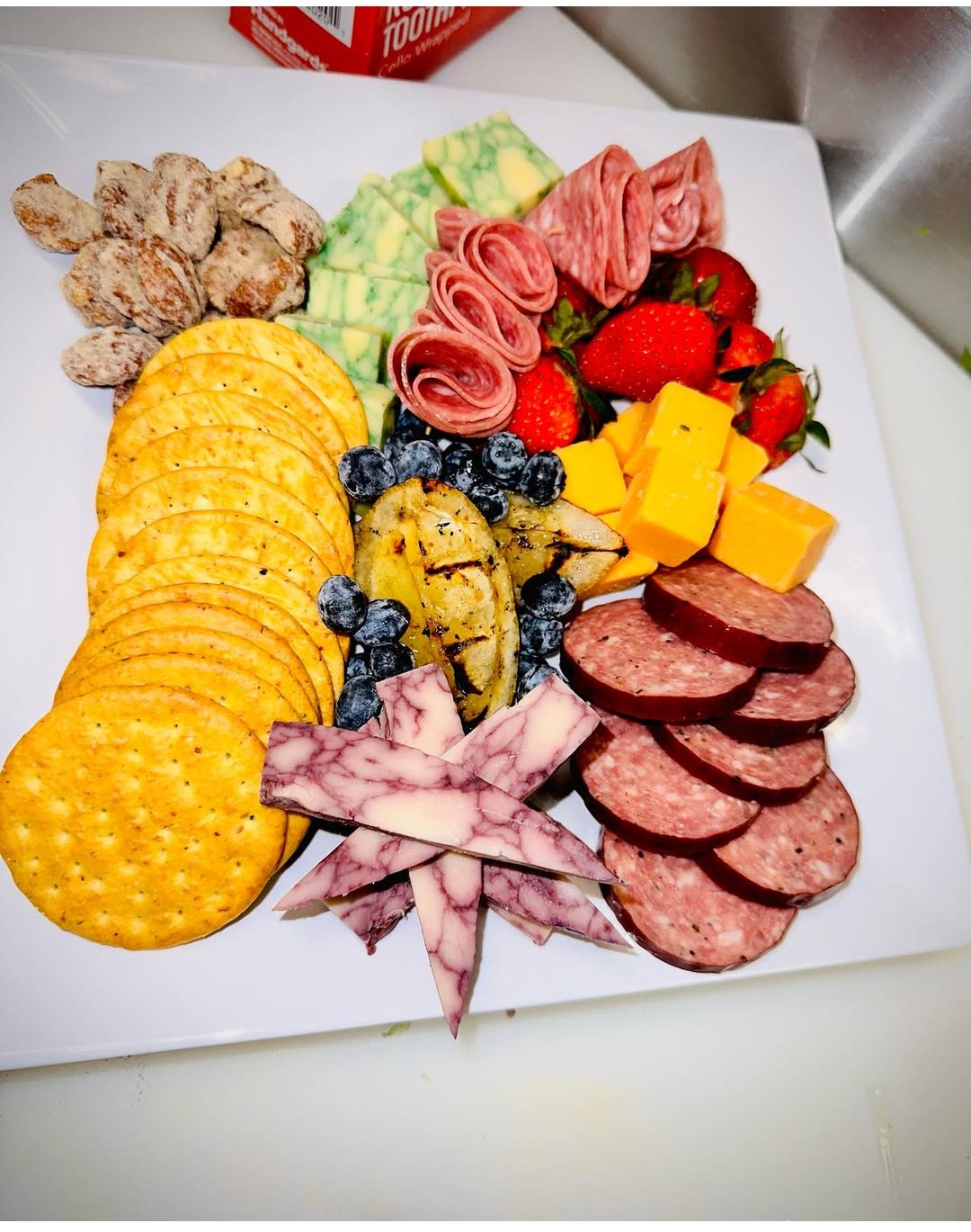 Assorted snacks including crackers, sliced meats, cheese, strawberries, blueberries, and sliced vegetables on a white plate.