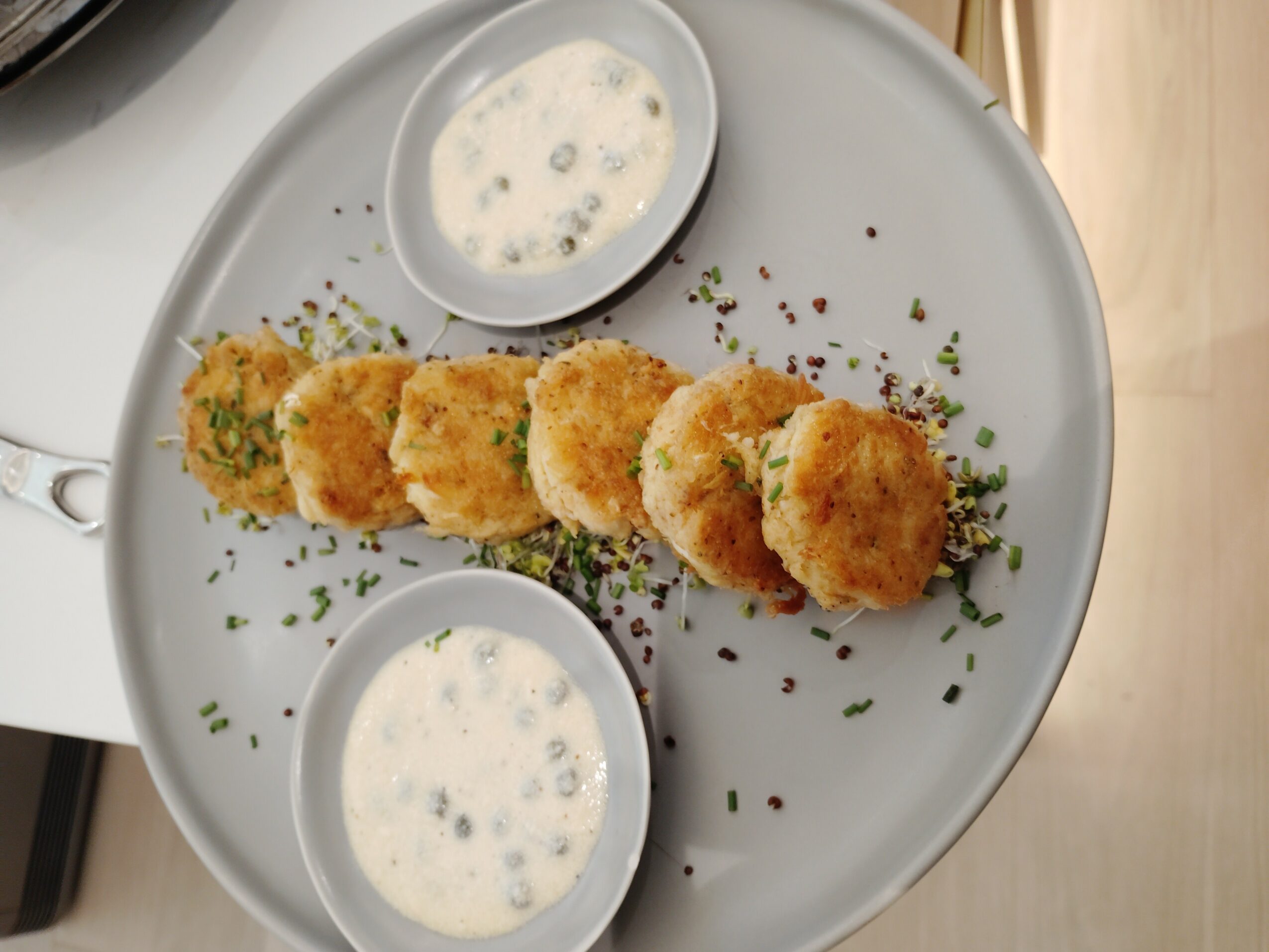 Plate with breaded fried food, two small bowls of white dipping sauce, garnished with herbs and pepper, on a white surface.