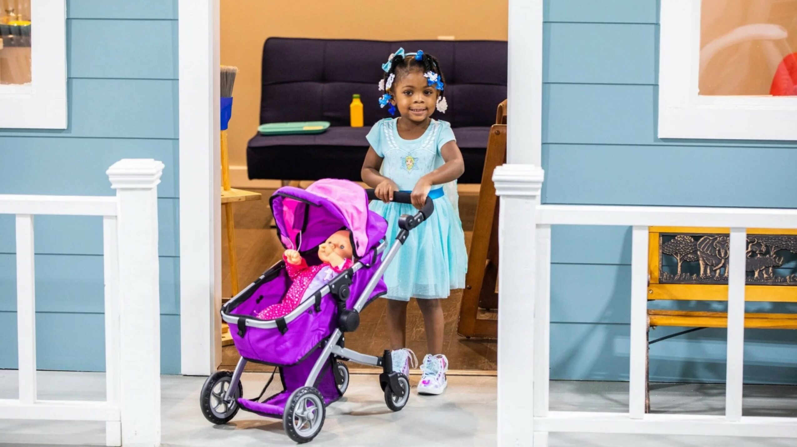 Child in a blue dress standing behind a purple stroller with a doll inside, in front of a blue house with white trim.