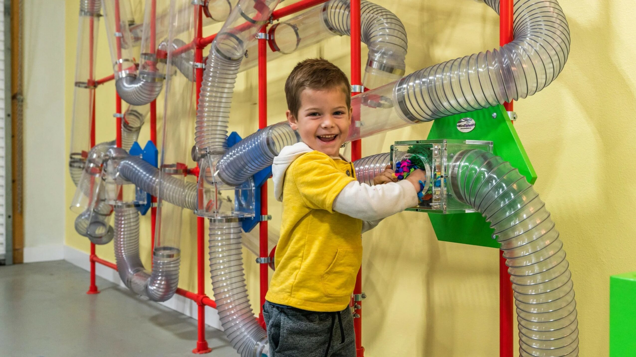 Child playing with large flexible tubes in an indoor play area, smiling and wearing a yellow shirt.