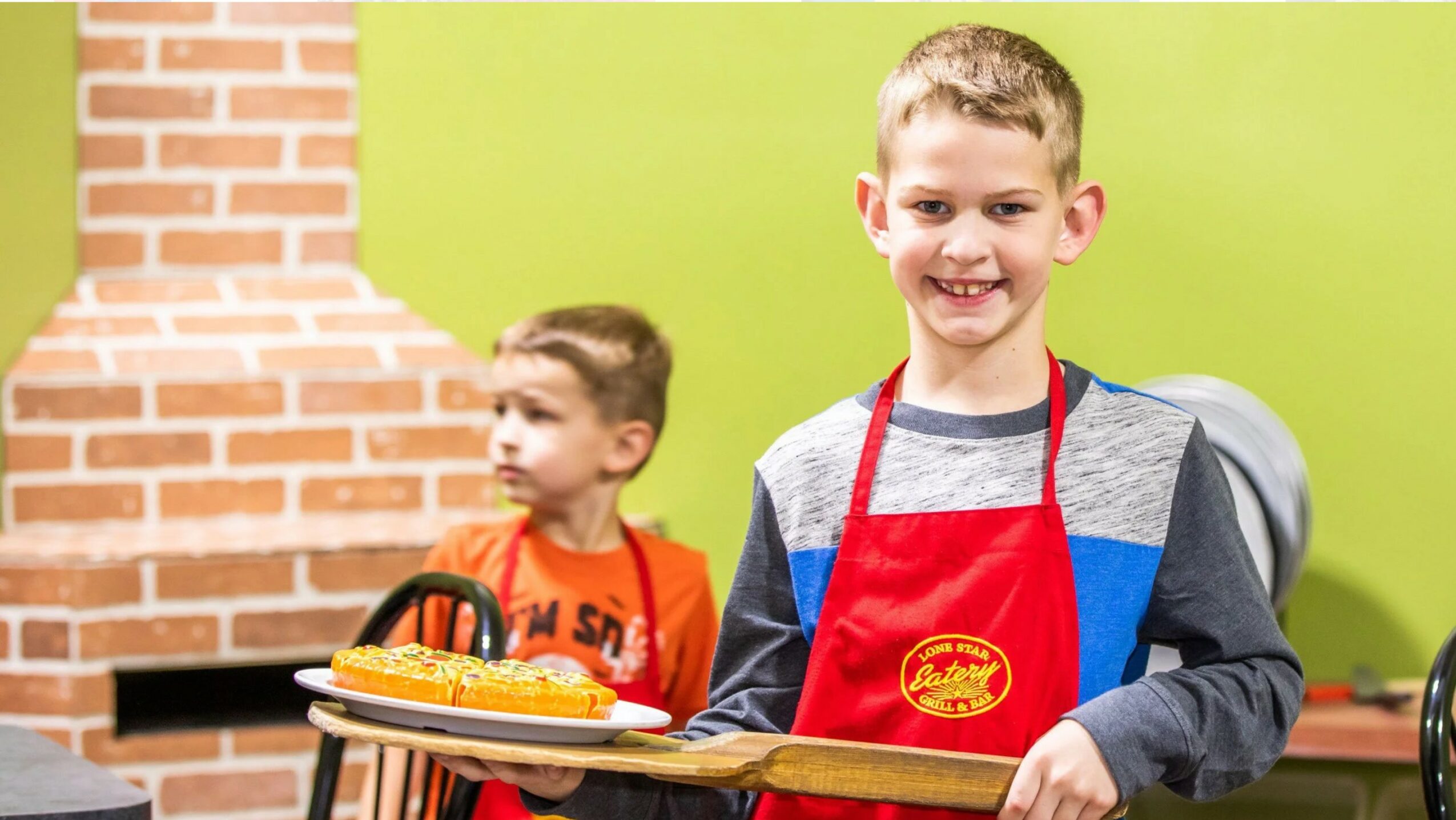 Two children in a kitchen, one smiling and wearing a red apron, holding a tray with food, brick wall background.