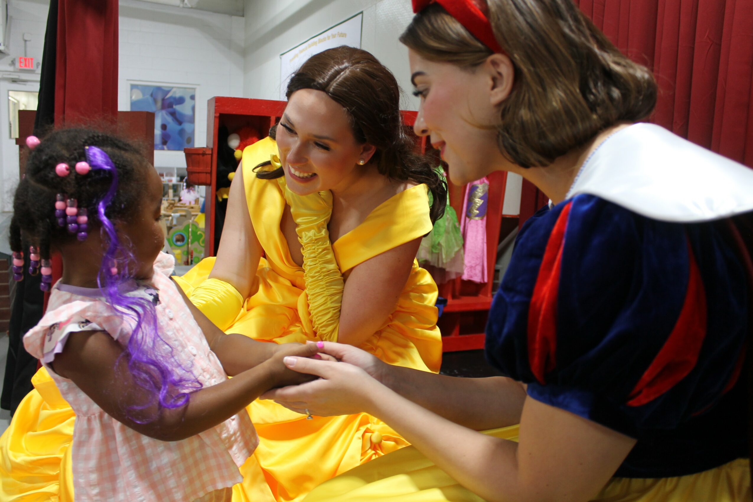 Two women and a girl holding hands and smiling indoors, with colorful decorations in the background.