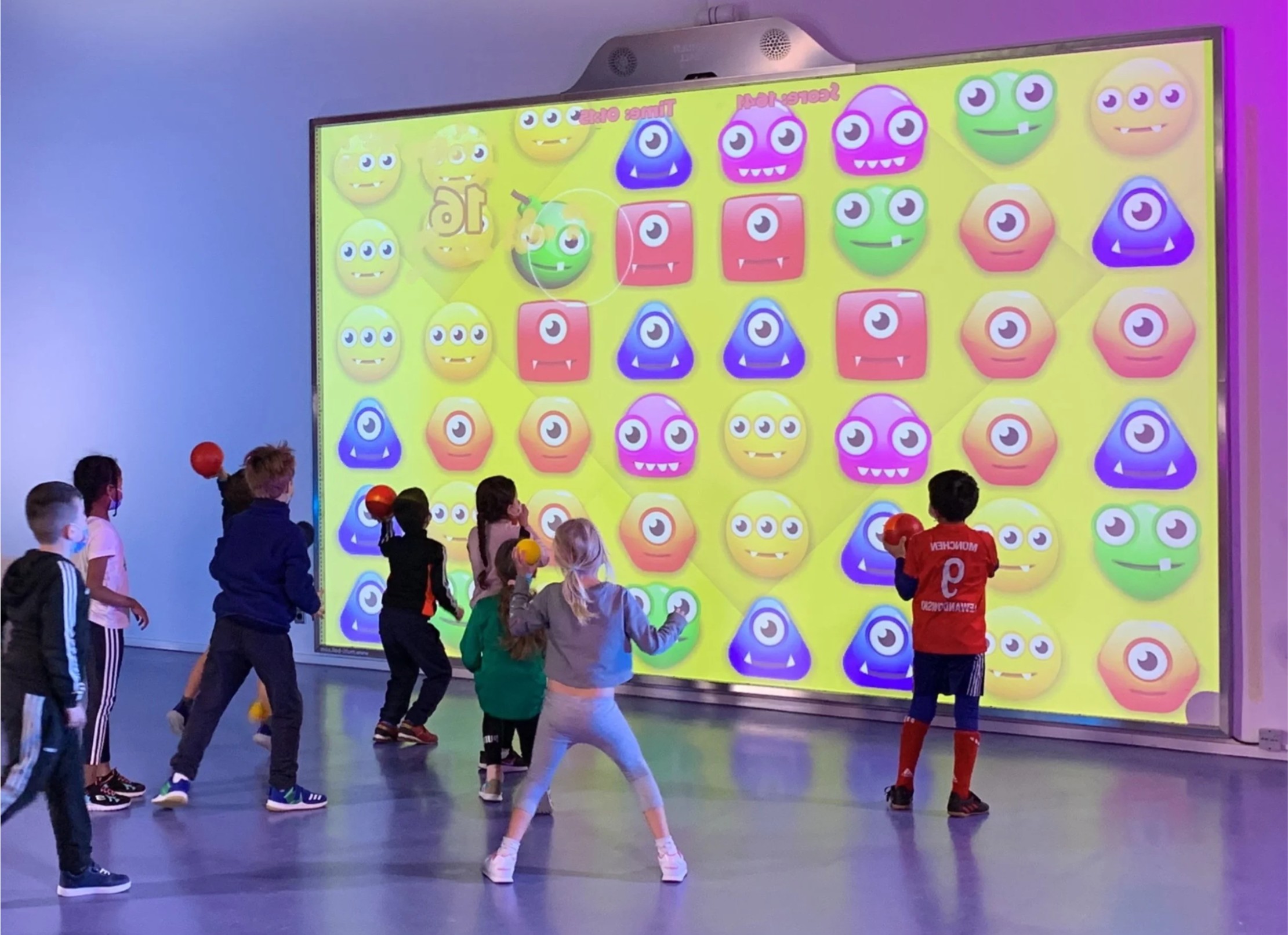 Children playing in front of a large colorful screen with smiley faces and geometric shapes, in a bright room.