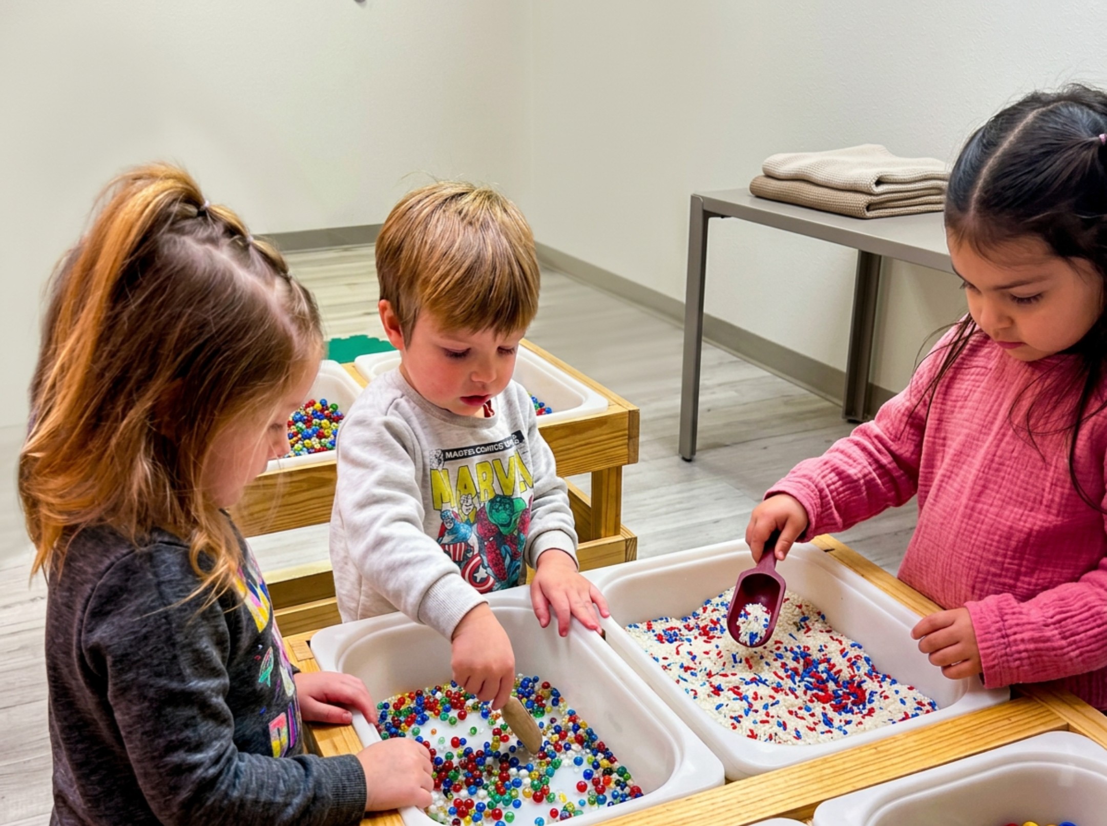 Three children playing with colorful beads in a room with a table and a towel on a stand.