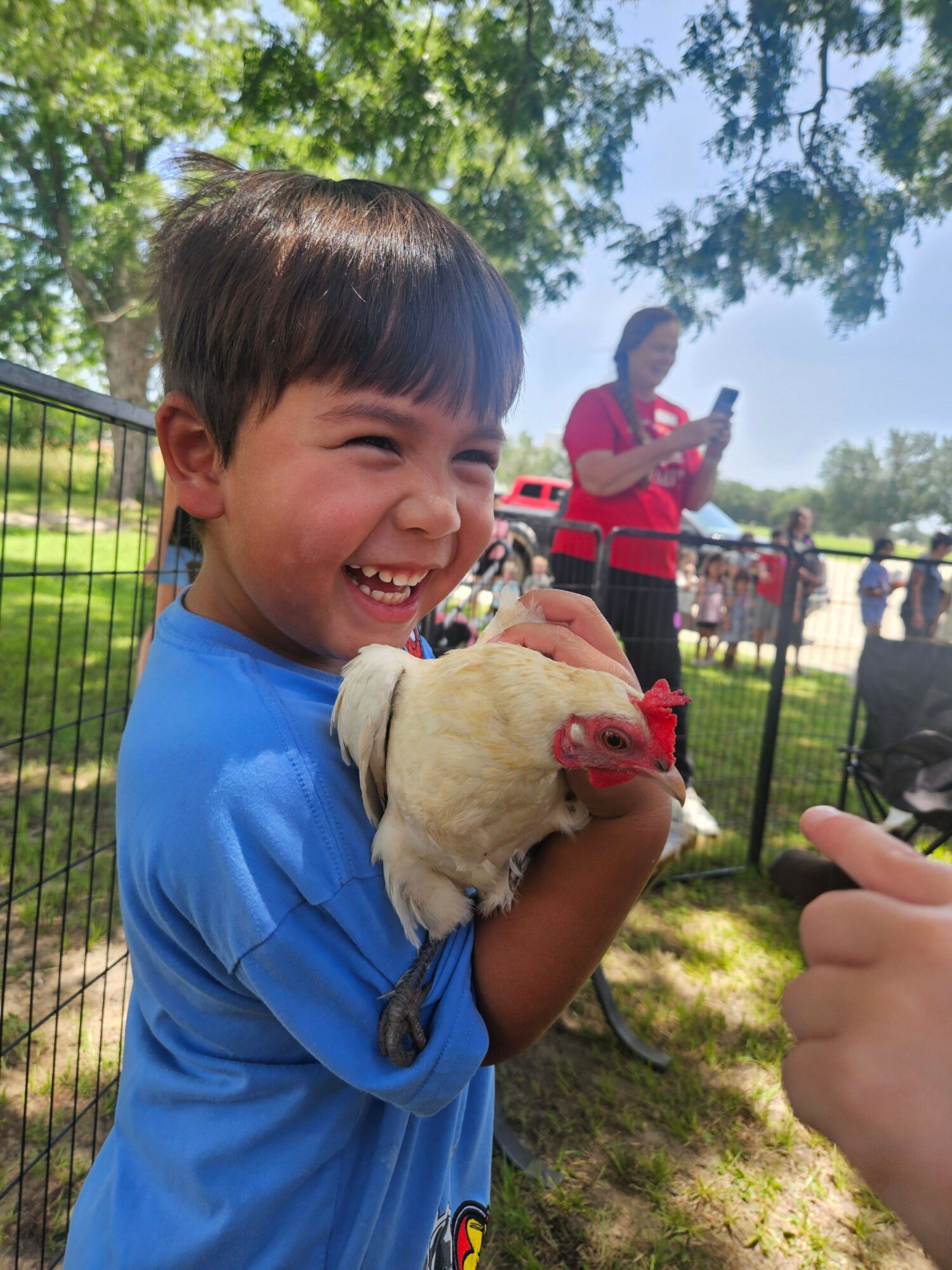 Child holding a chicken outdoors, smiling, with people and trees in the background.