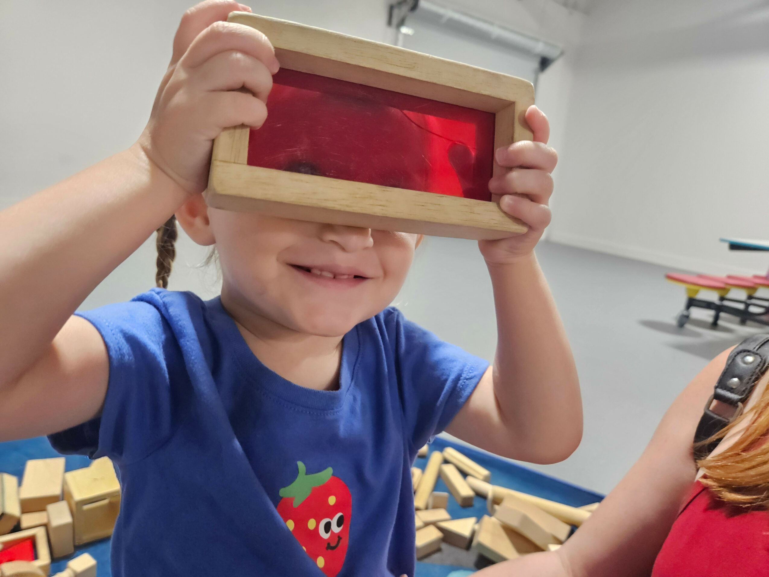 Child holding a wooden frame over eyes, smiling, with wooden blocks scattered around, in a classroom setting.