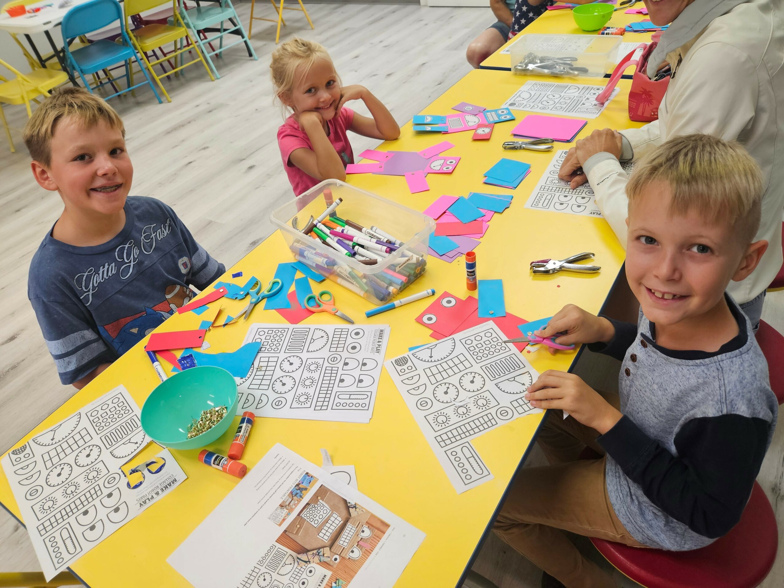 Children sit at a yellow table with colorful paper, markers, and worksheets, engaging in arts and crafts activities.