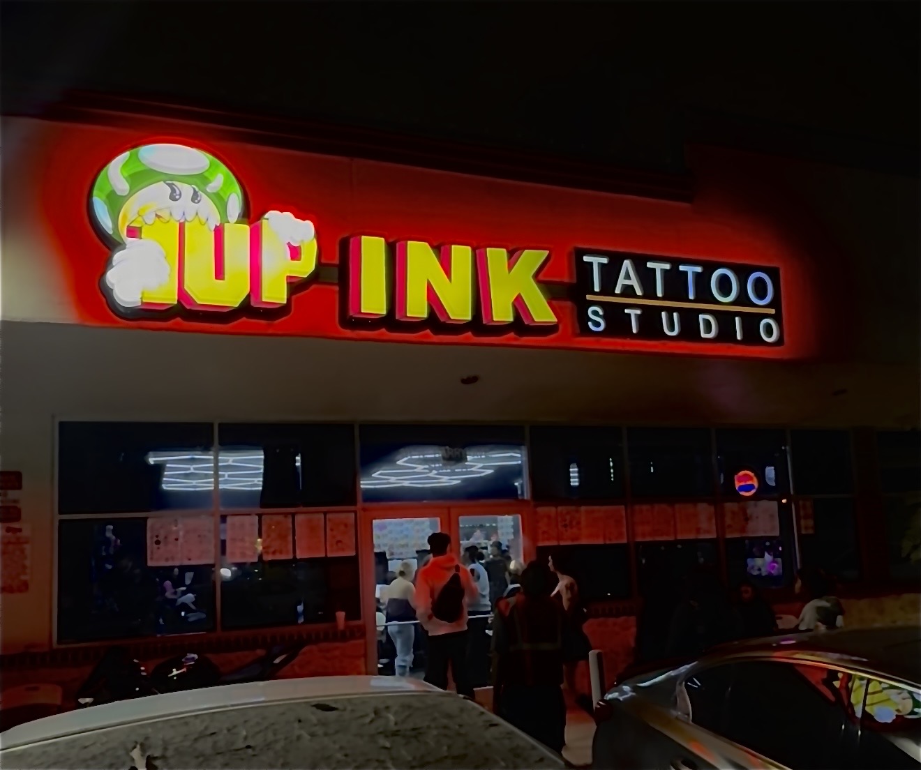 Bright illuminated sign for tattoo studio with people outside, cars parked in front, and a dark background.
