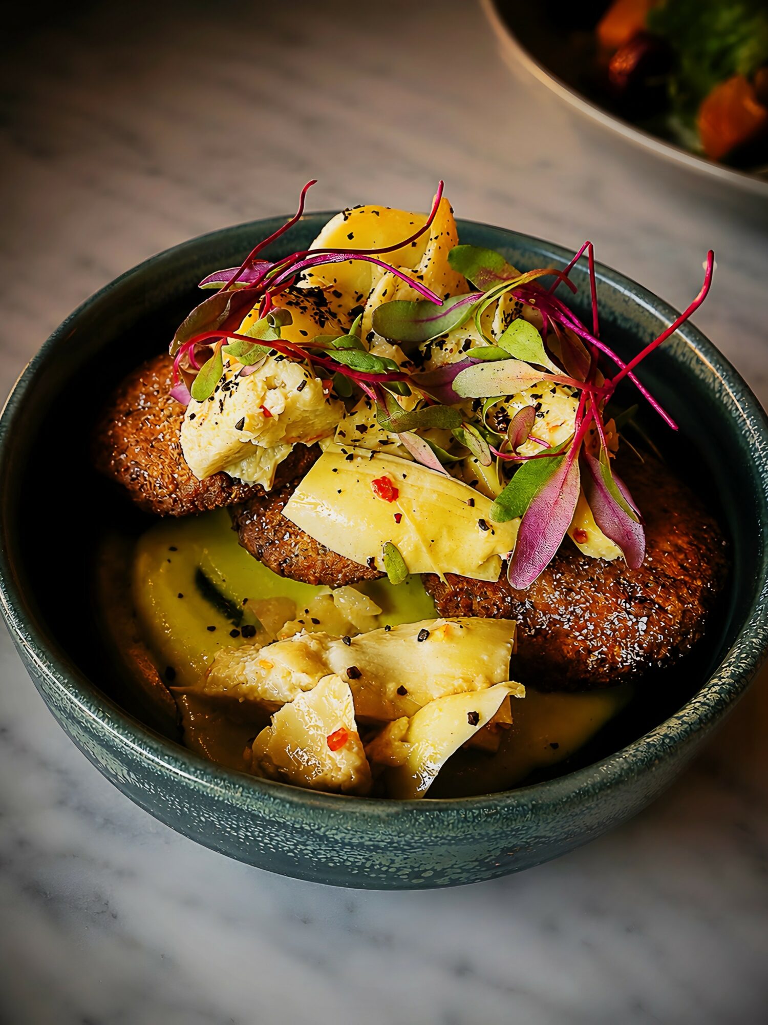 A bowl of food with sliced potatoes, greens, and garnishes, presented on a dark surface.