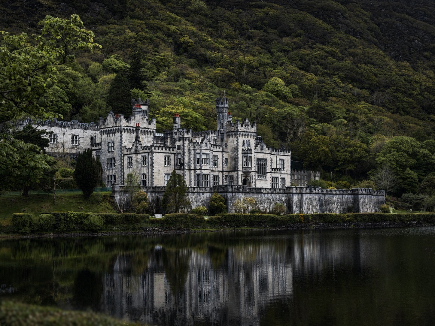 Castle with towers and walls beside a lake, surrounded by green trees and hillside, reflected in the water.