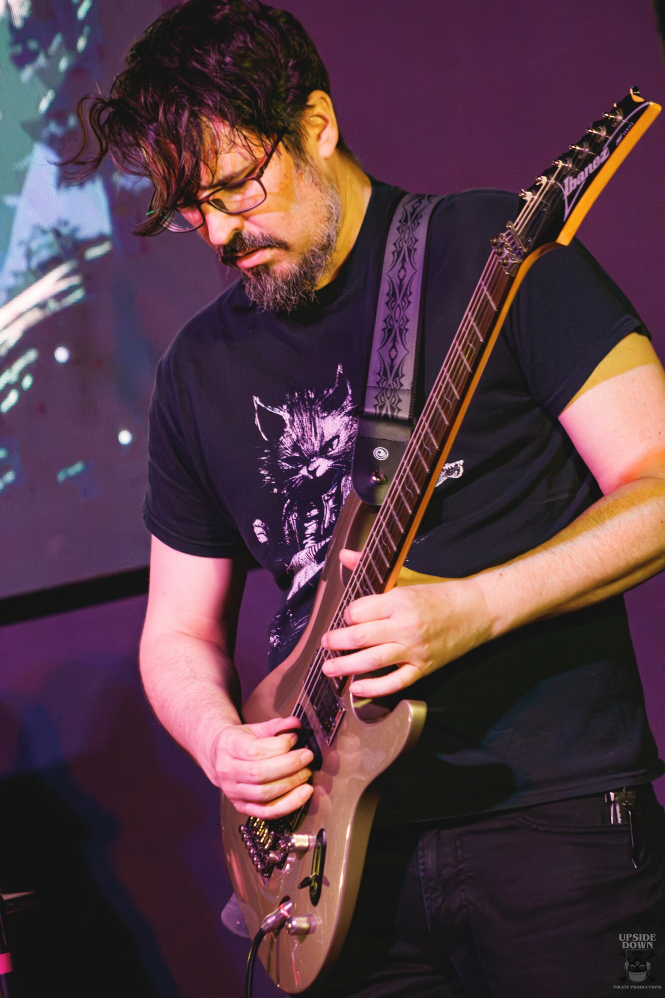 Person with curly hair and glasses playing an electric guitar on stage, wearing a black t-shirt with a graphic design.