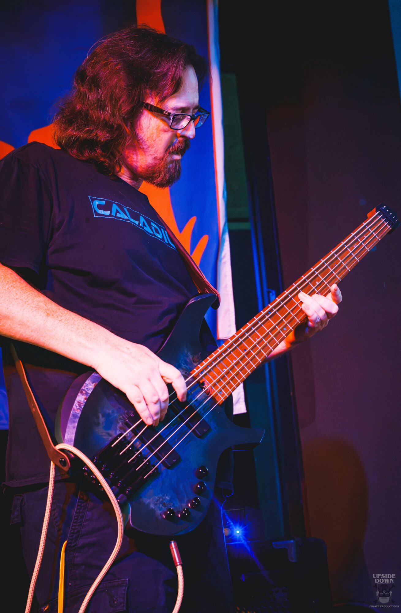 Person with long hair and glasses playing a black electric bass guitar on stage, with colorful background lighting.