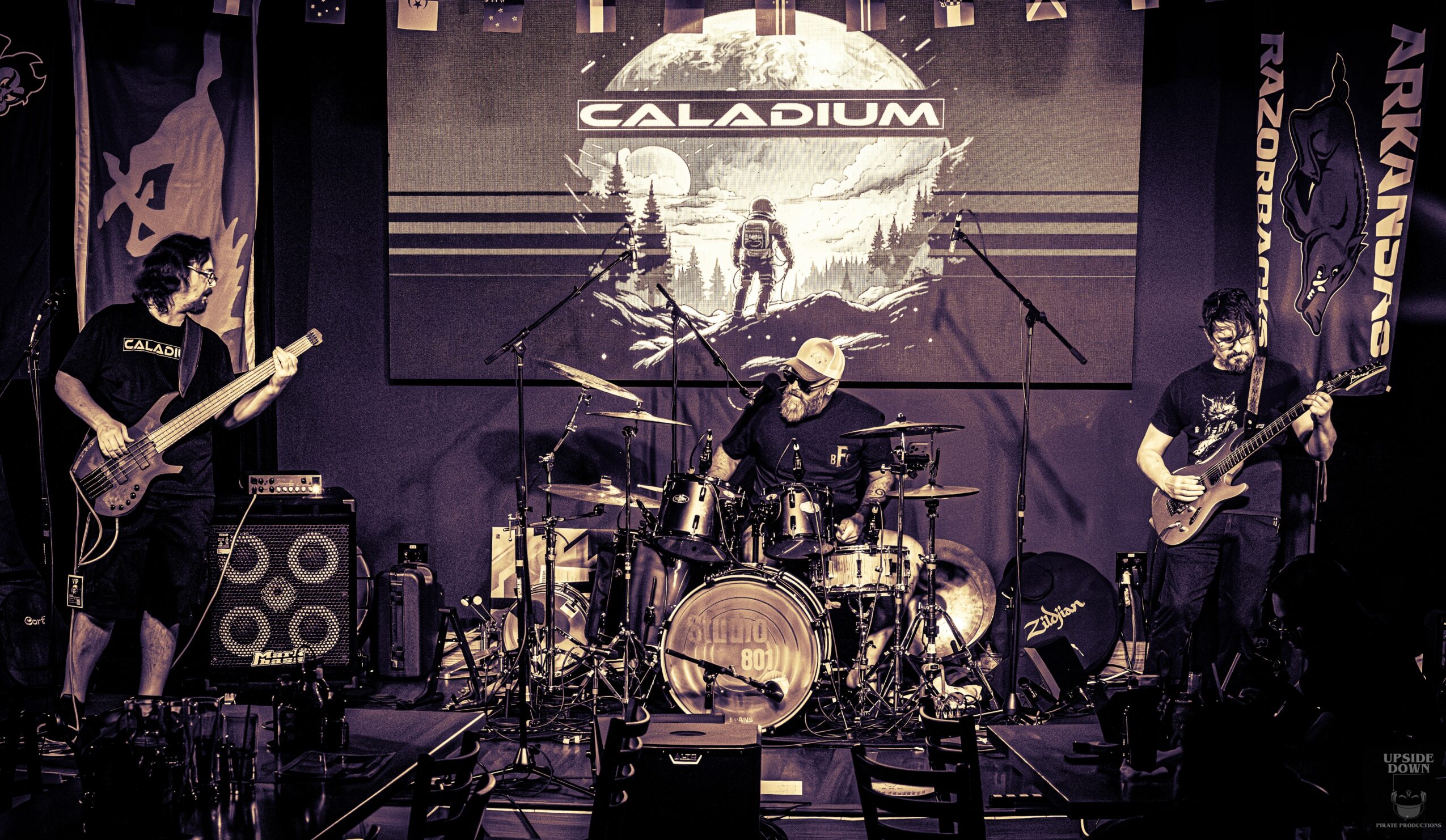 Three musicians perform on stage with guitars and drums, a screen displays a logo and landscape behind them.