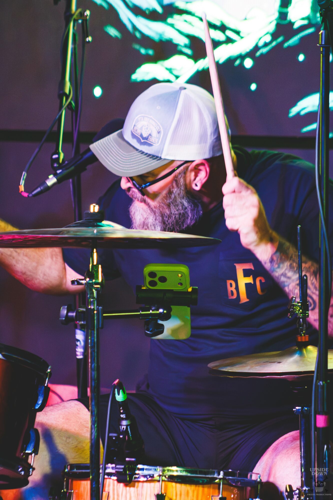 A drummer wearing a white cap and glasses playing drums on stage with colorful background lights.
