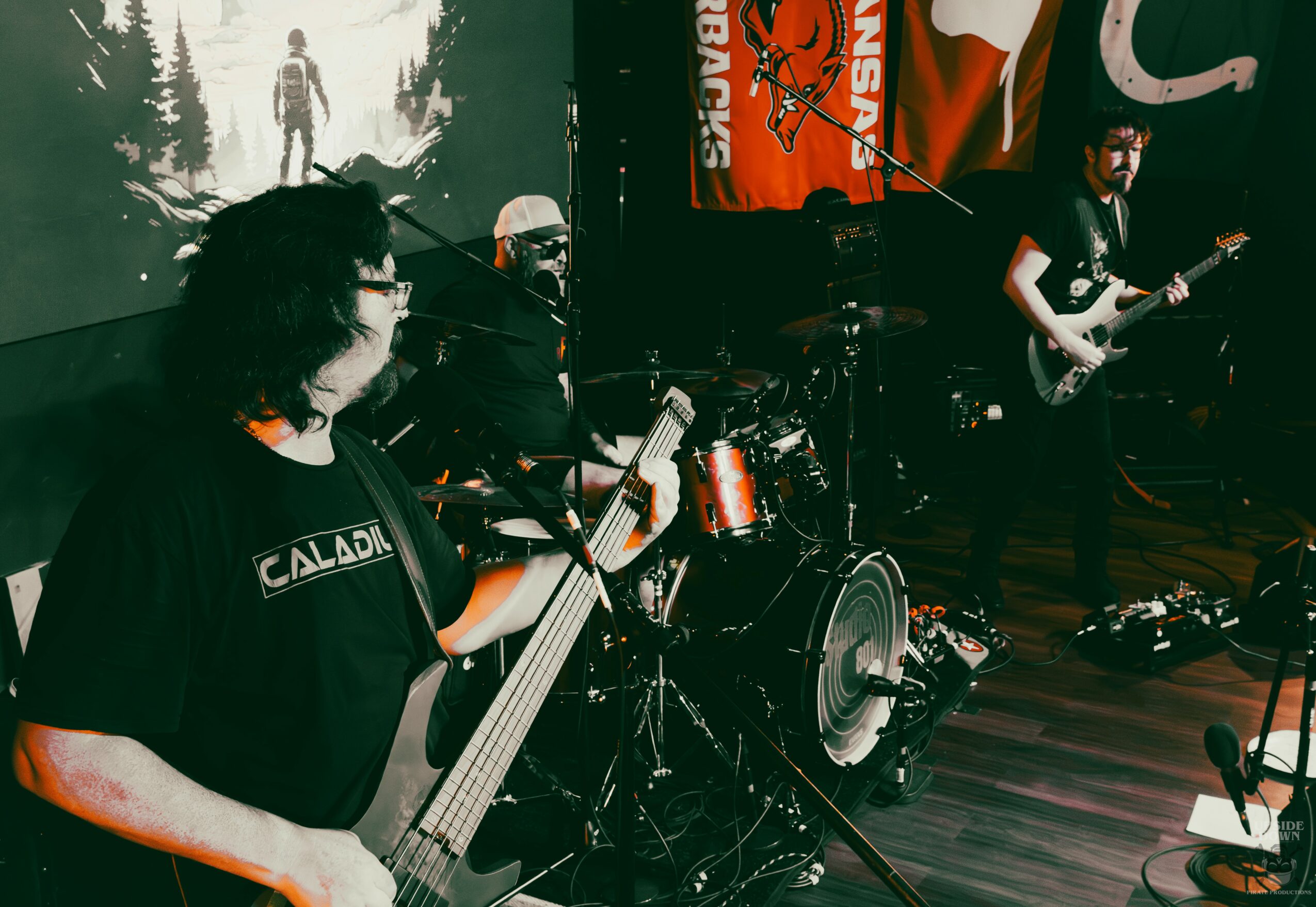 Band performing on stage with three musicians playing guitars and drums, flags and a forest scene projected in background.
