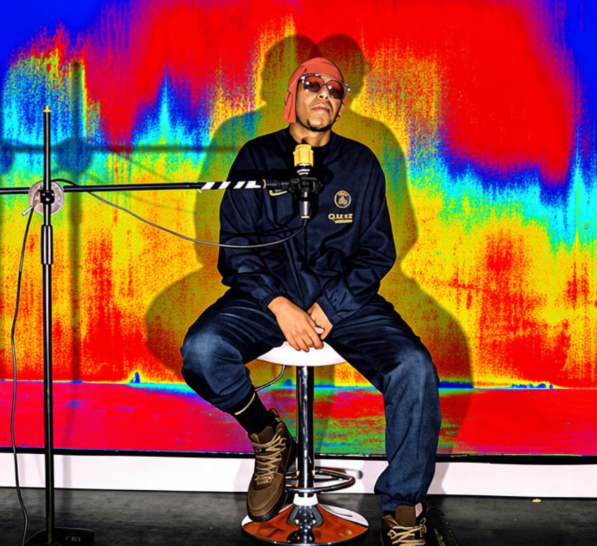Person sitting on a stool with a colorful, abstract background, wearing sunglasses and a dark jacket, in a studio setting.
