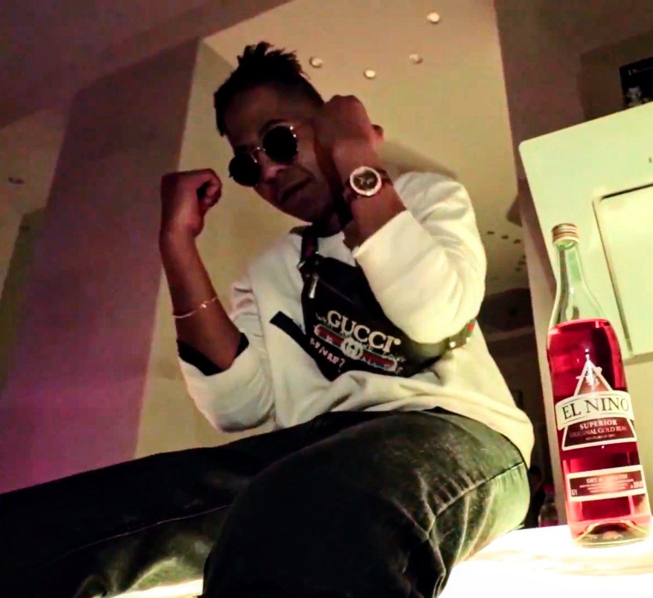 Person wearing sunglasses and a white Gucci sweatshirt, sitting with arms raised, in an indoor setting.