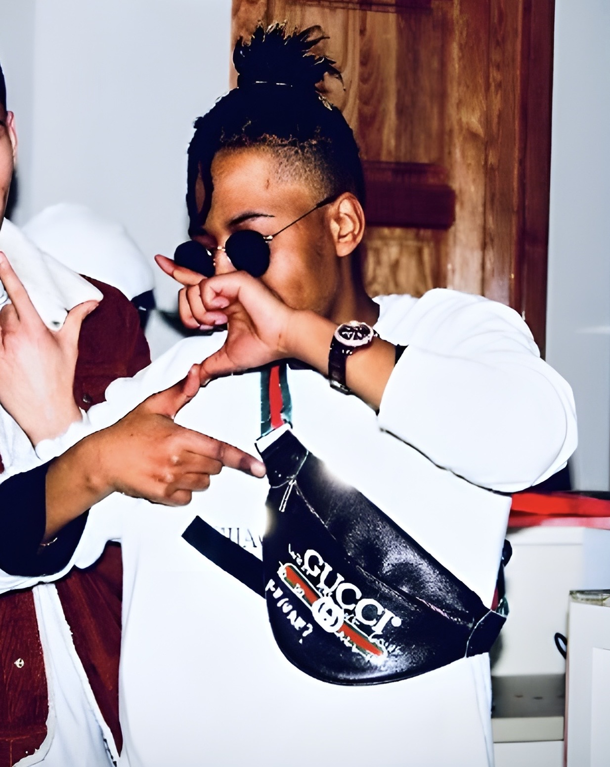 Person with sunglasses and a white jacket, pointing at a black Gucci bag, in an indoor setting.