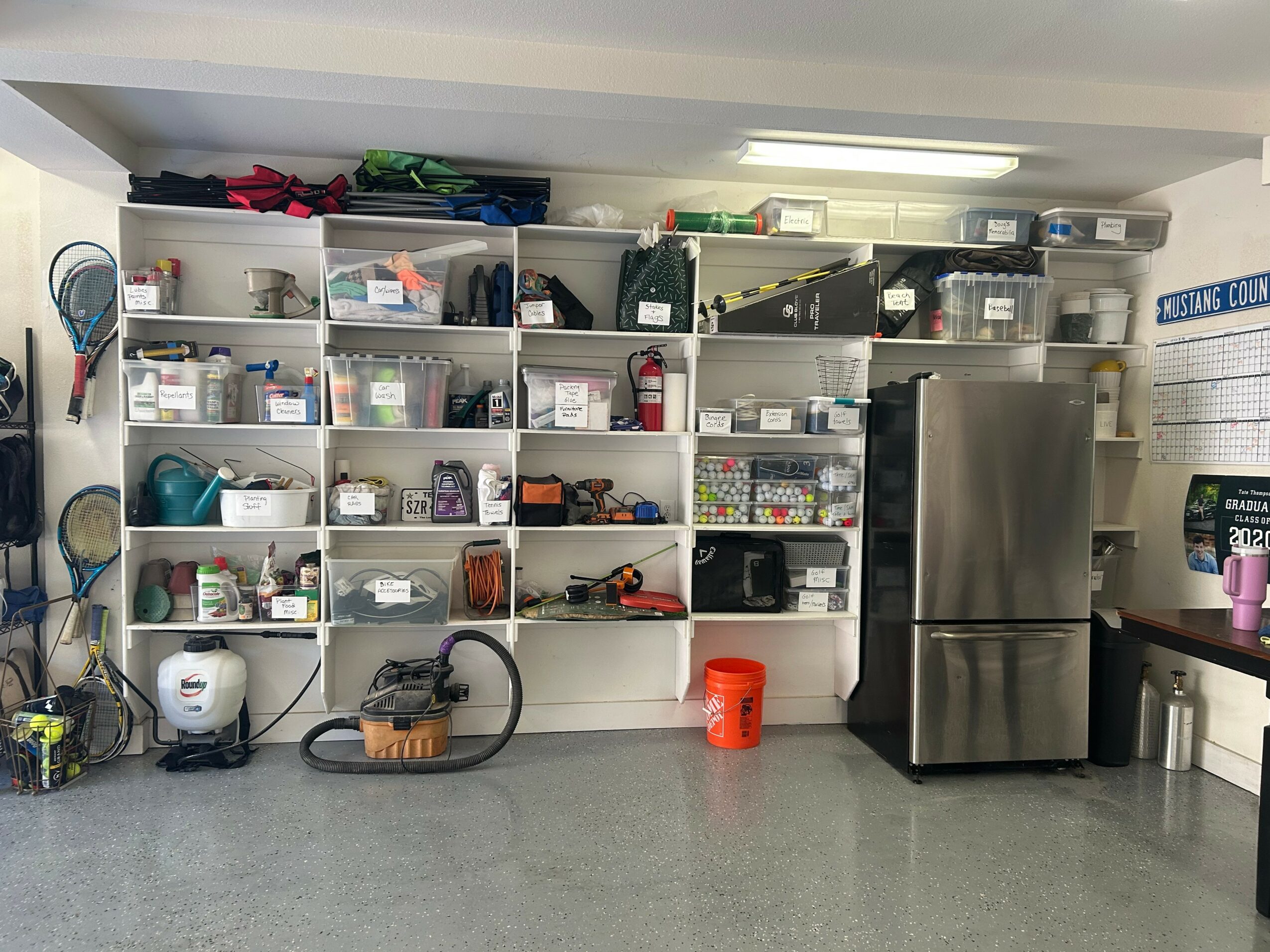 Shelves filled with various household items, a stainless steel refrigerator, and a small orange bucket on the floor.