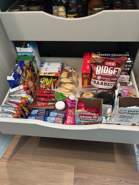 Open drawer filled with snacks, chips, and packaged food items, with a wooden floor visible below.