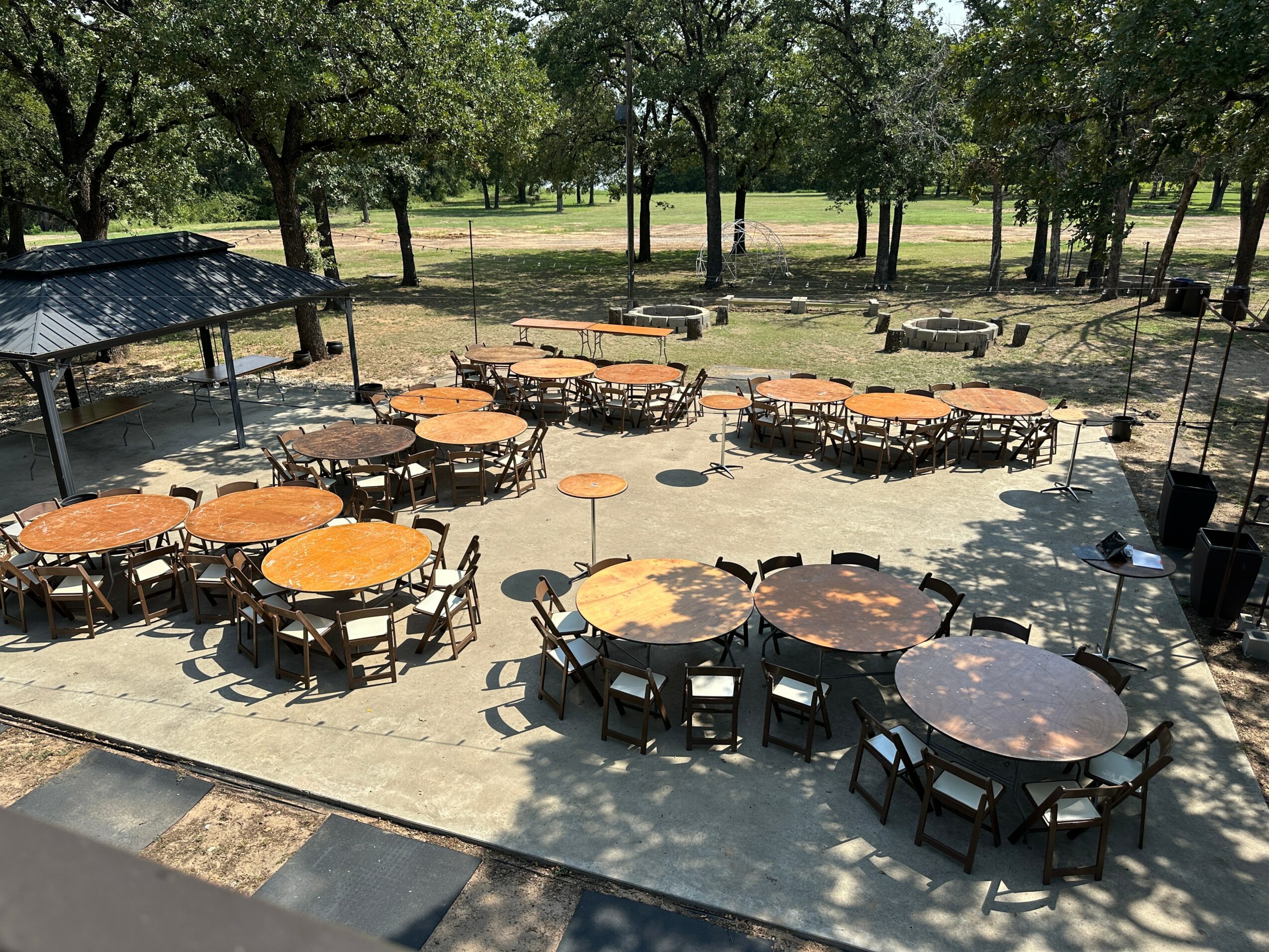 Outdoor seating area with round tables and chairs on a concrete surface, surrounded by trees and a park, sunny day.