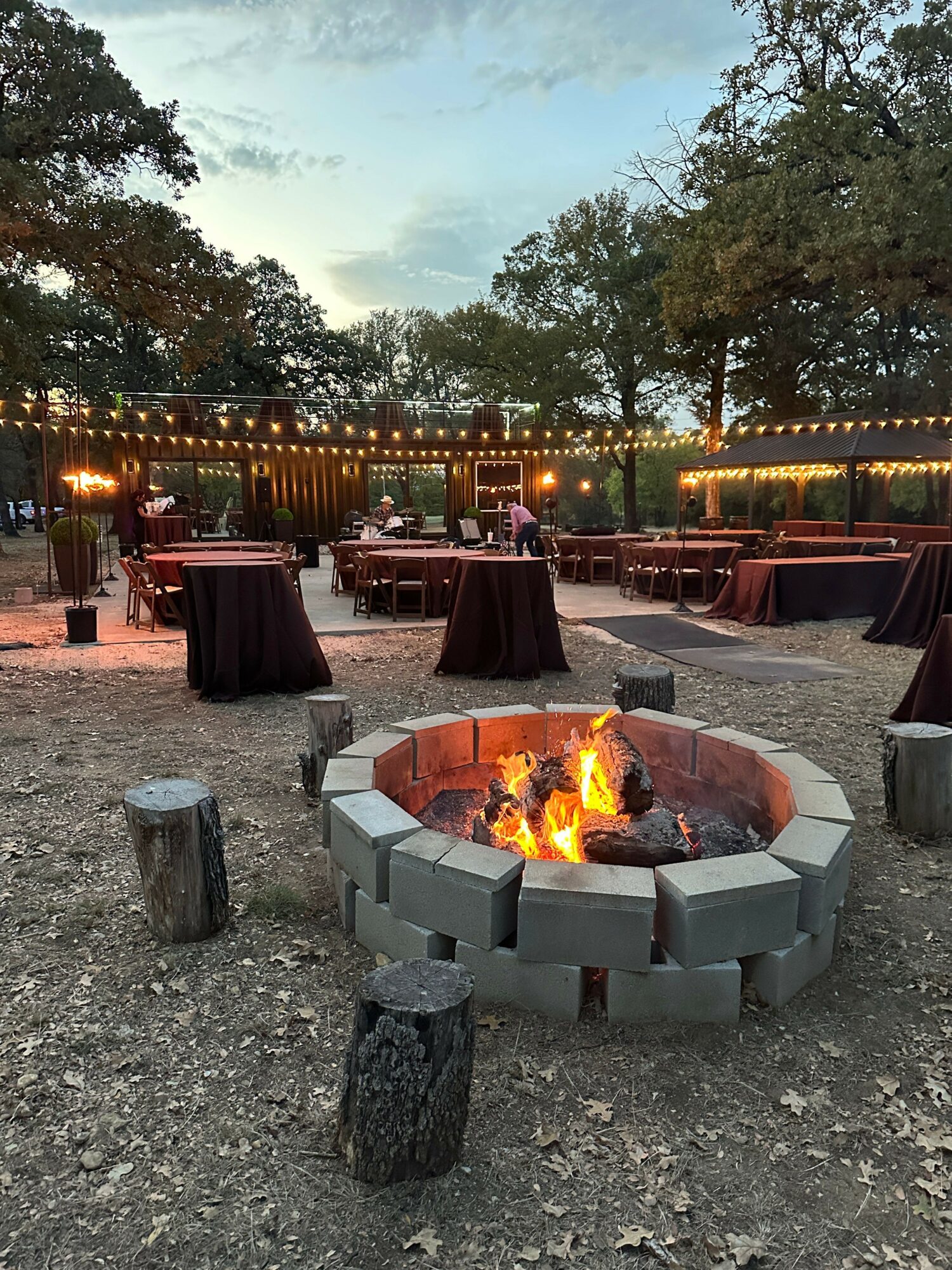 Outdoor fire pit surrounded by chairs and tables, with string lights and trees in the background during evening.