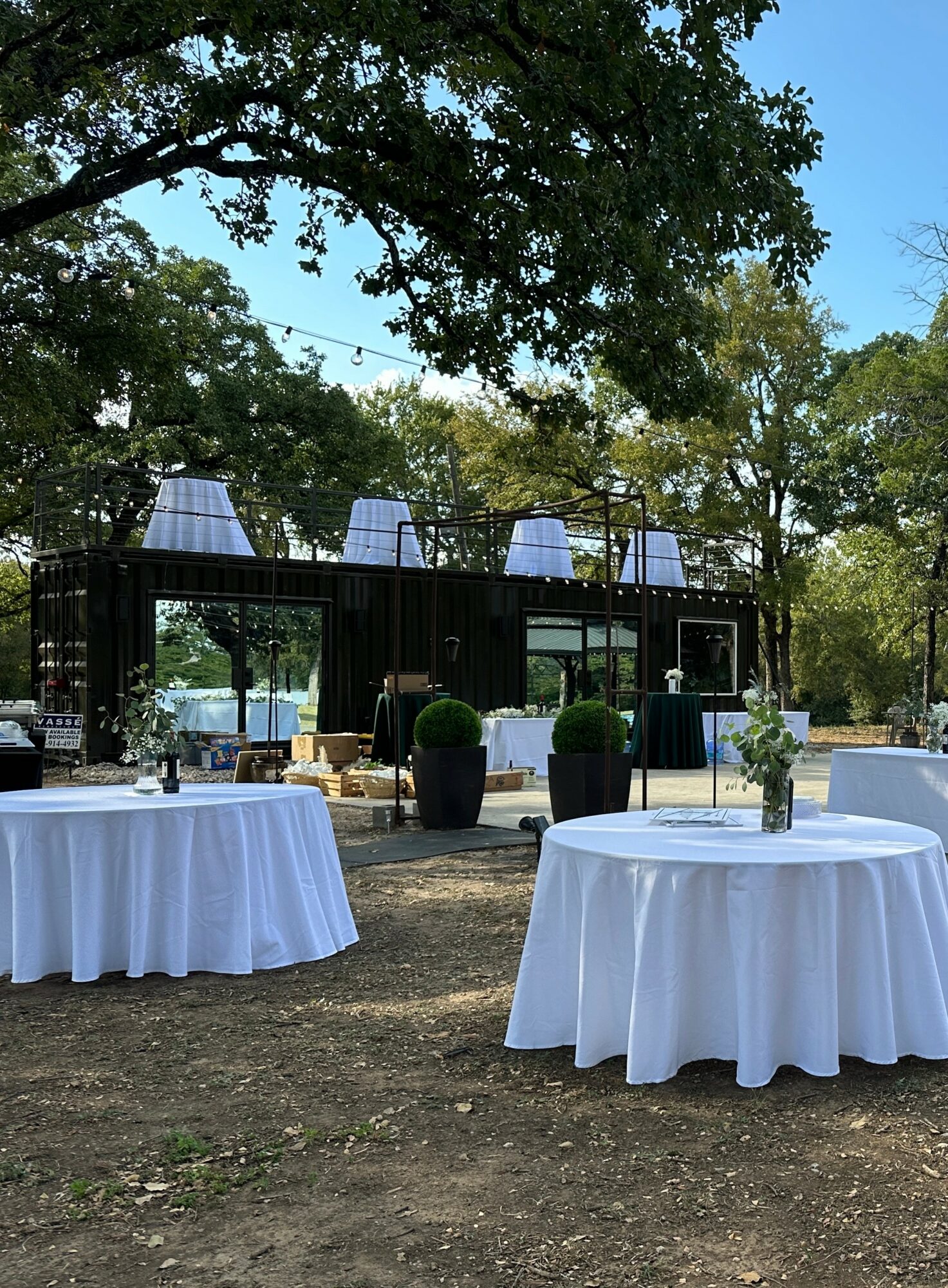 Outdoor event setup with round tables covered in white tablecloths and a black stage with lighting, surrounded by trees.