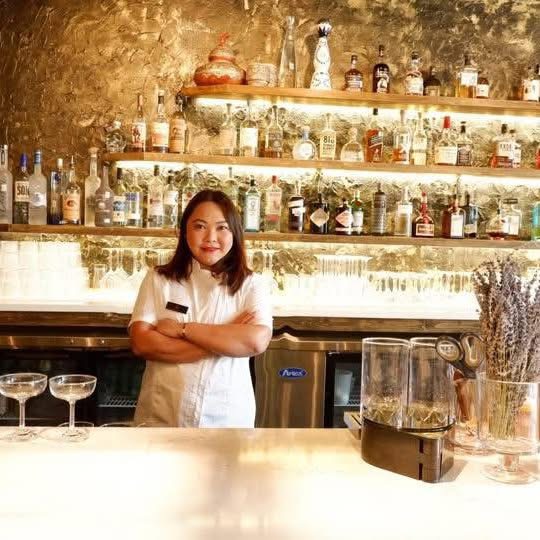 Woman standing behind a bar with shelves of bottles and jars, arms crossed, smiling.