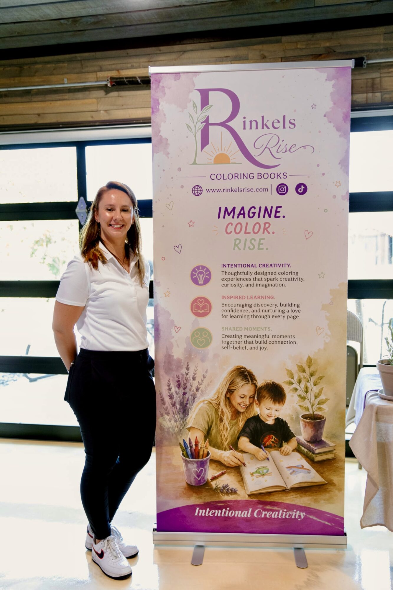 Woman standing next to a vertical banner with purple and pink design, children drawing at a table, and text about coloring books.