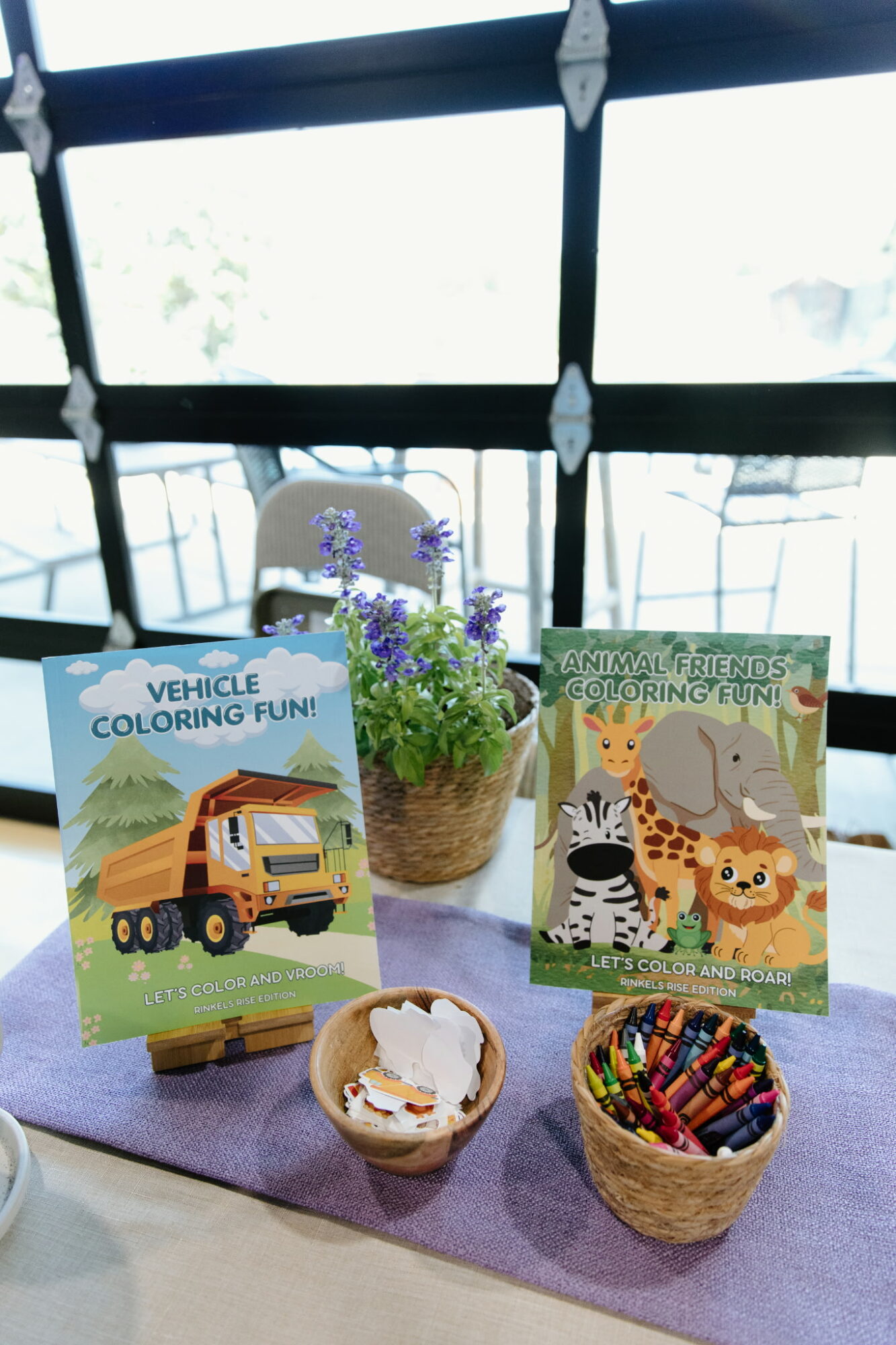 Table with potted plant, animal-themed coloring books, and baskets of crayons, set near a window with a grid pattern.