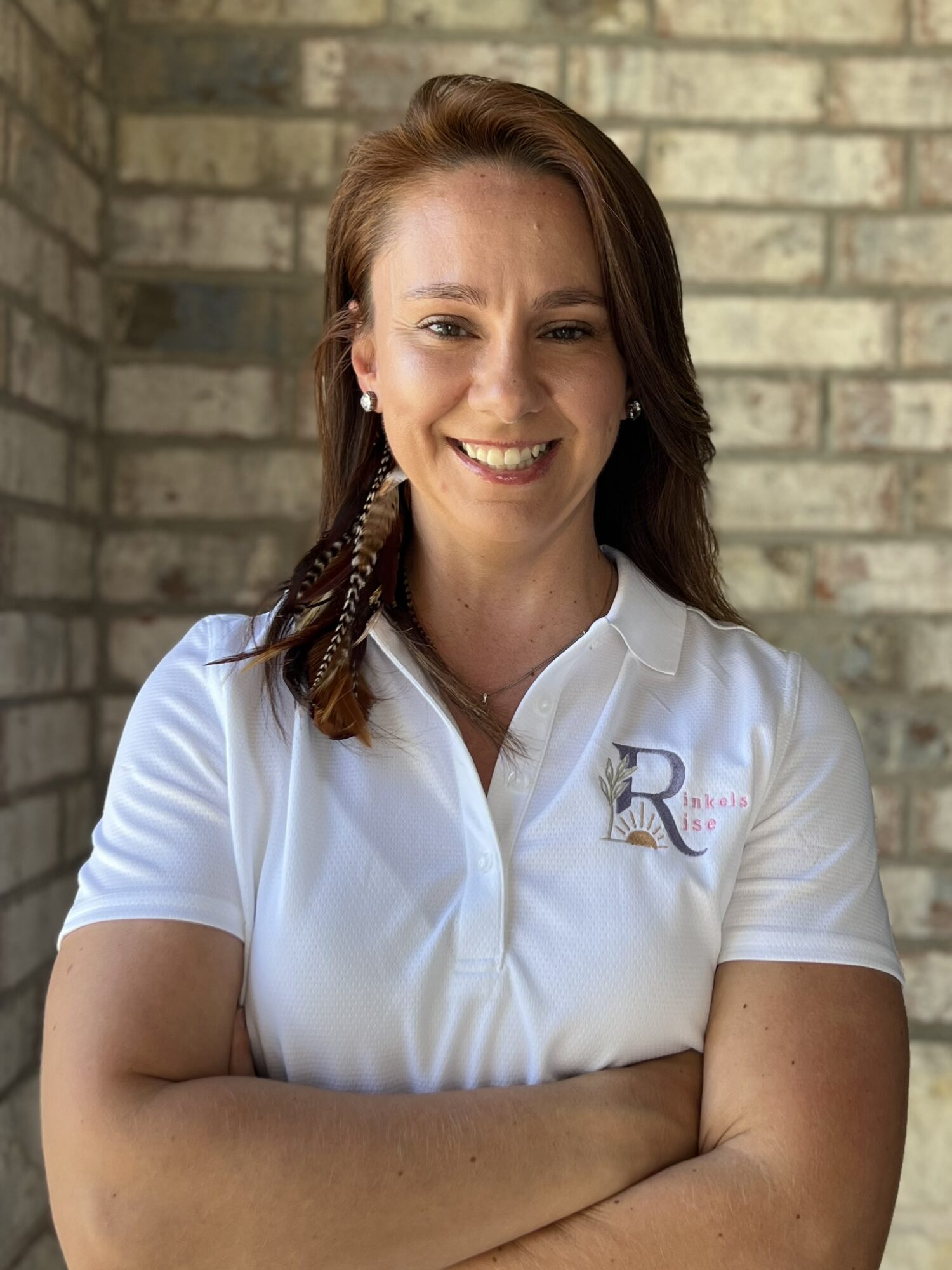Woman with long brown hair smiling, wearing a white polo shirt with a logo, standing in front of a brick wall.