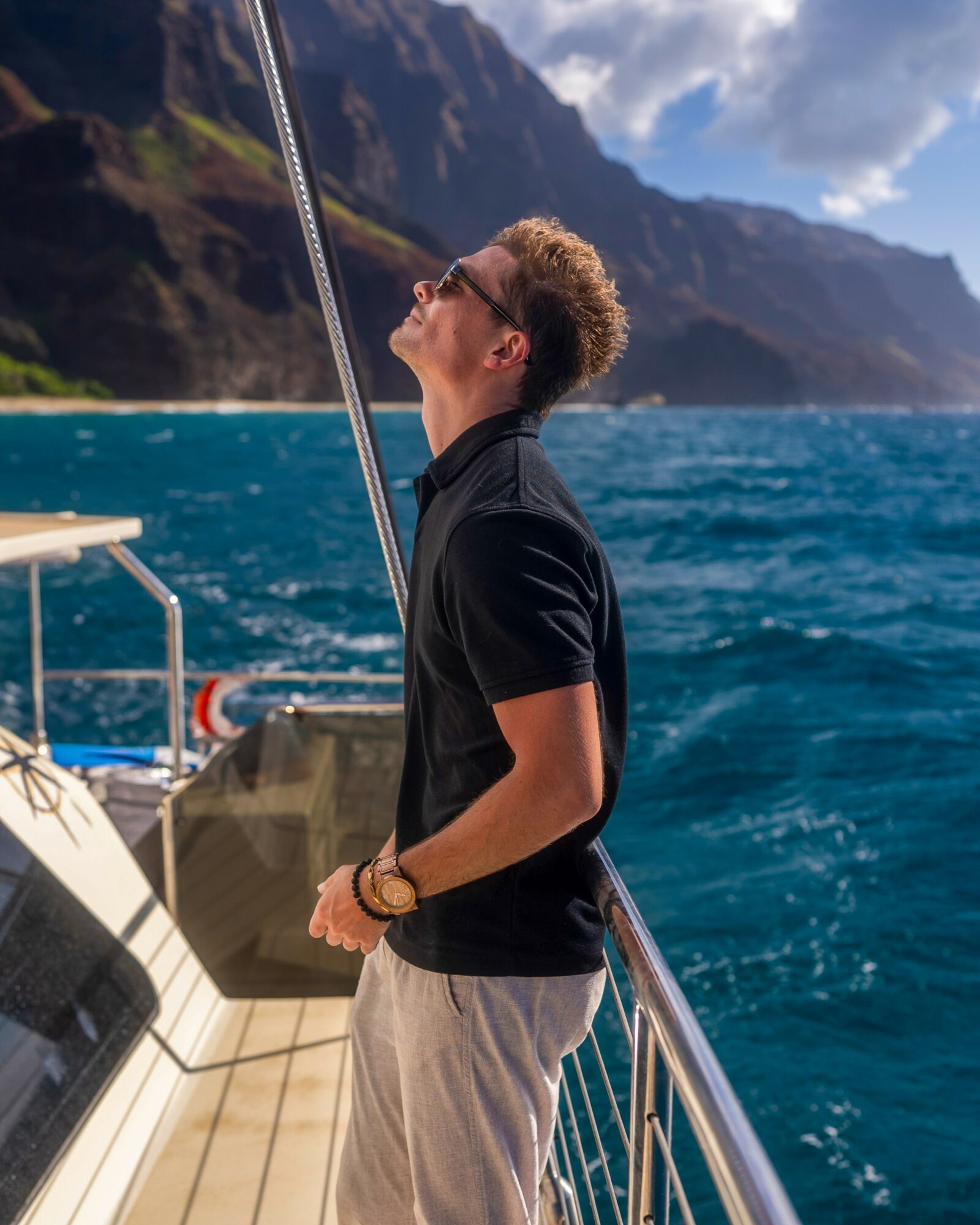 Young man with curly hair wearing sunglasses, black polo shirt, and beige pants, standing on a boat railing, looking up at the sky.