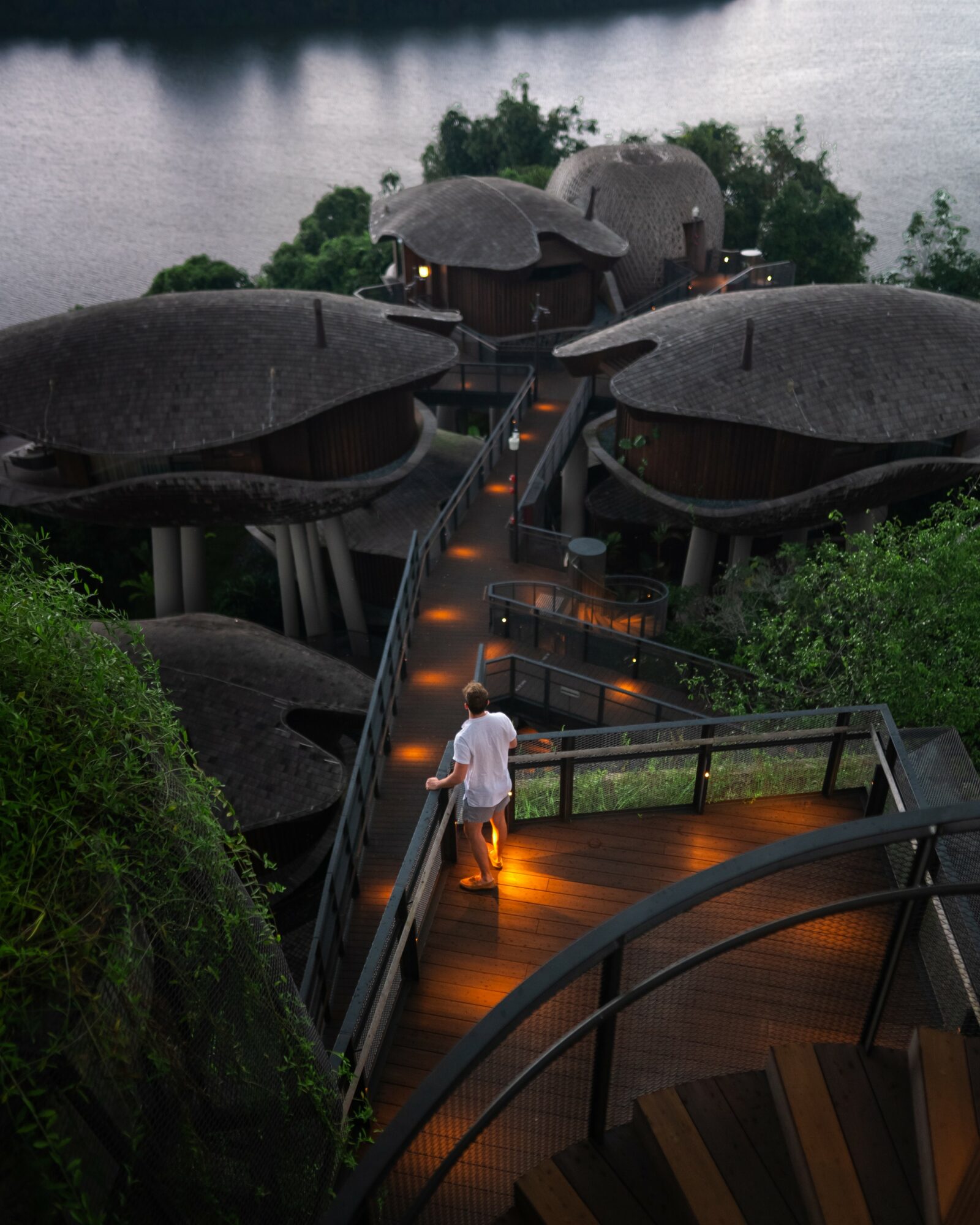 Person walking on a wooden bridge towards unique treehouse-style structures over water, surrounded by greenery at night.