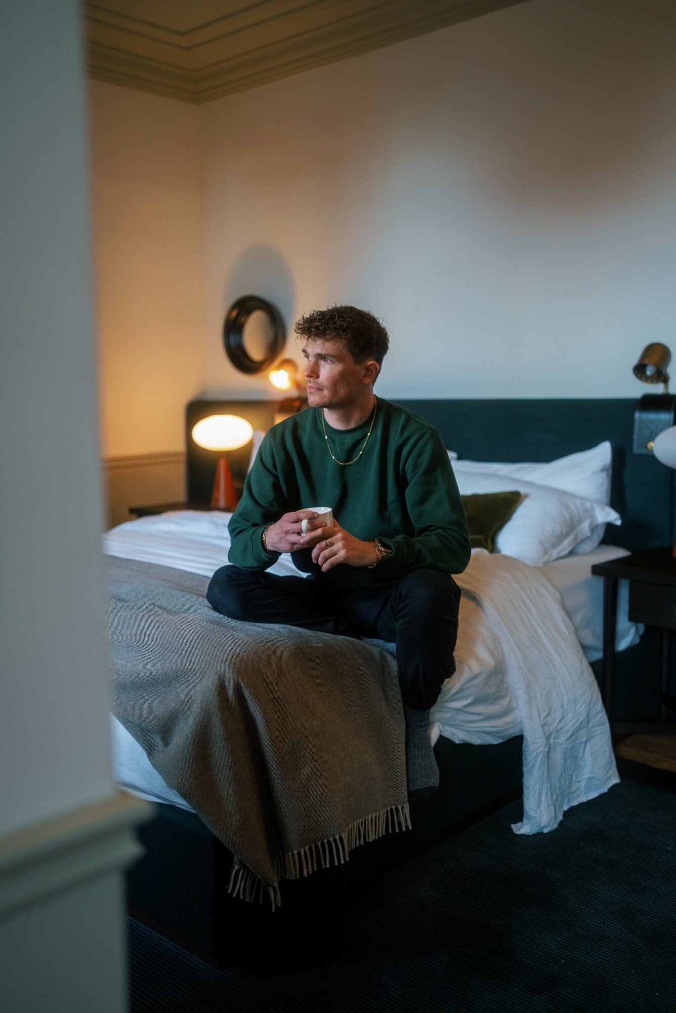 Person sitting on a bed in a dimly lit room, holding a cup, with lamps and decor in the background.