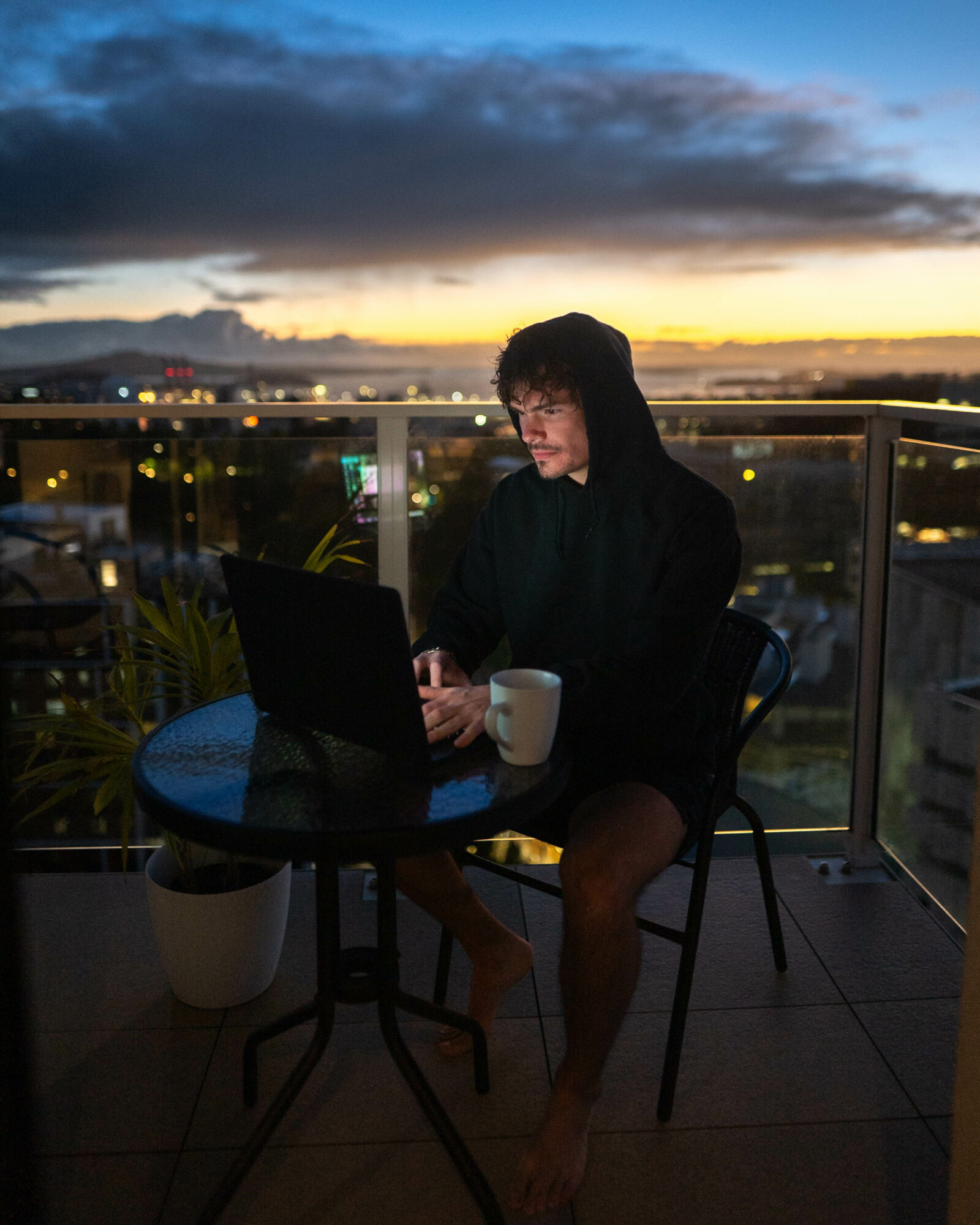 Person working on a laptop at a table on a balcony during sunset, with a cityscape in the background.