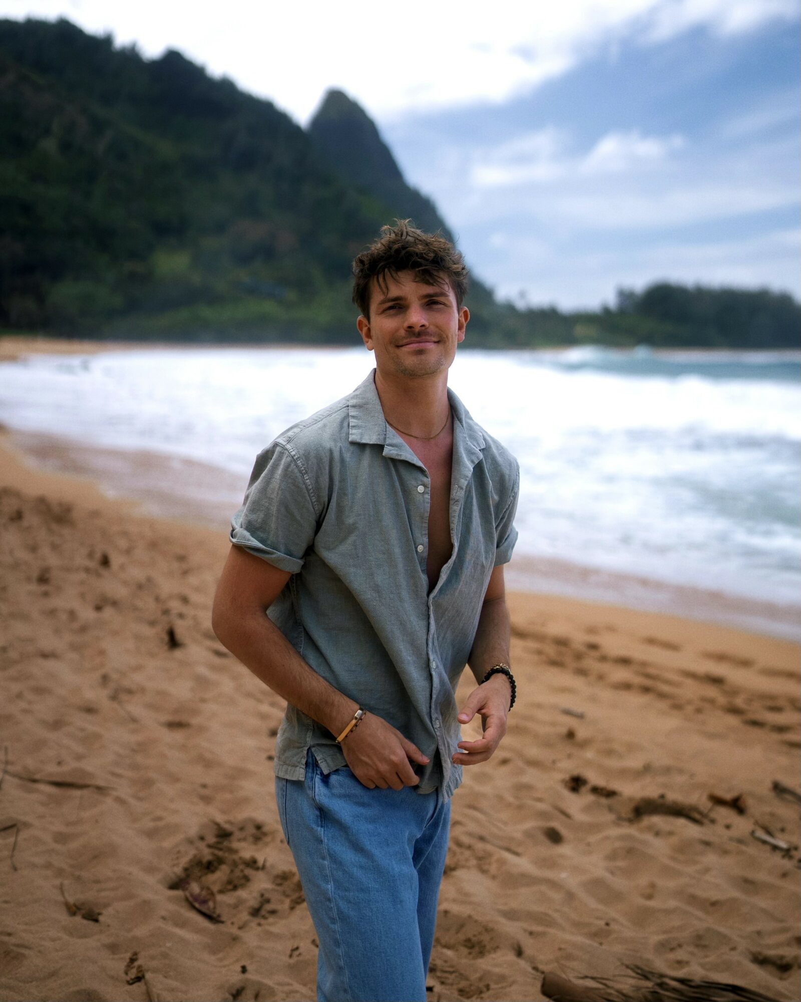 Young man standing on a sandy beach with ocean waves and green mountains in the background, smiling at the camera.