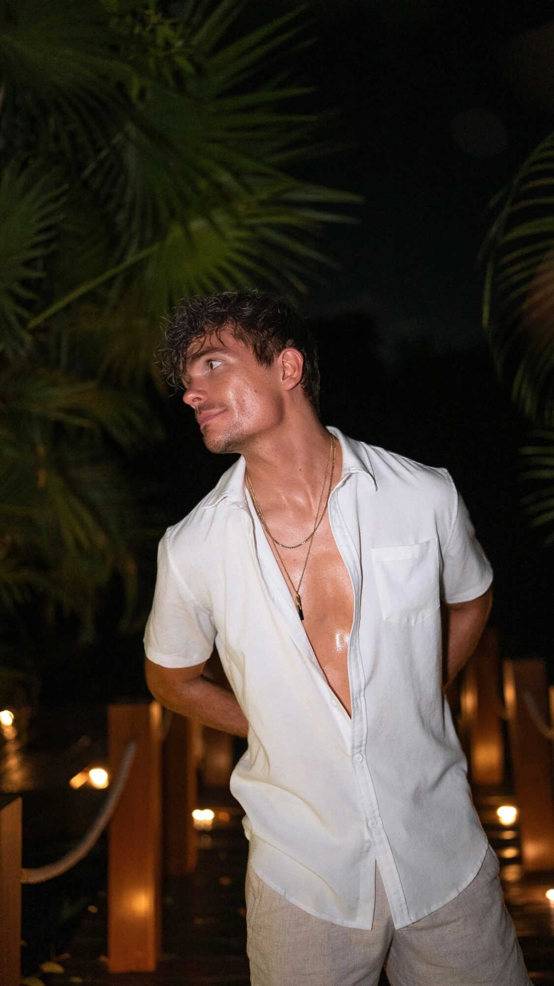 Young man with short hair and a necklace, wearing a partially unbuttoned white shirt, standing outdoors at night among plants.