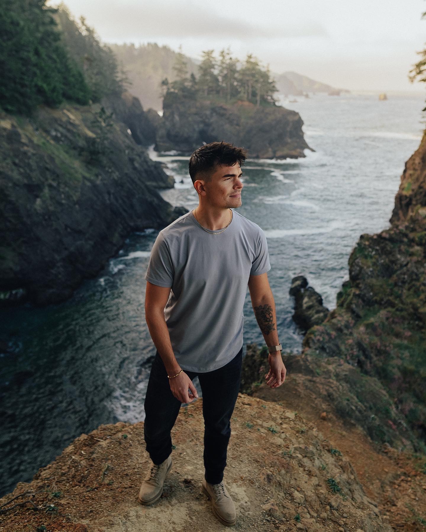 Young man standing on rocky terrain near water with cliffs and trees in background.