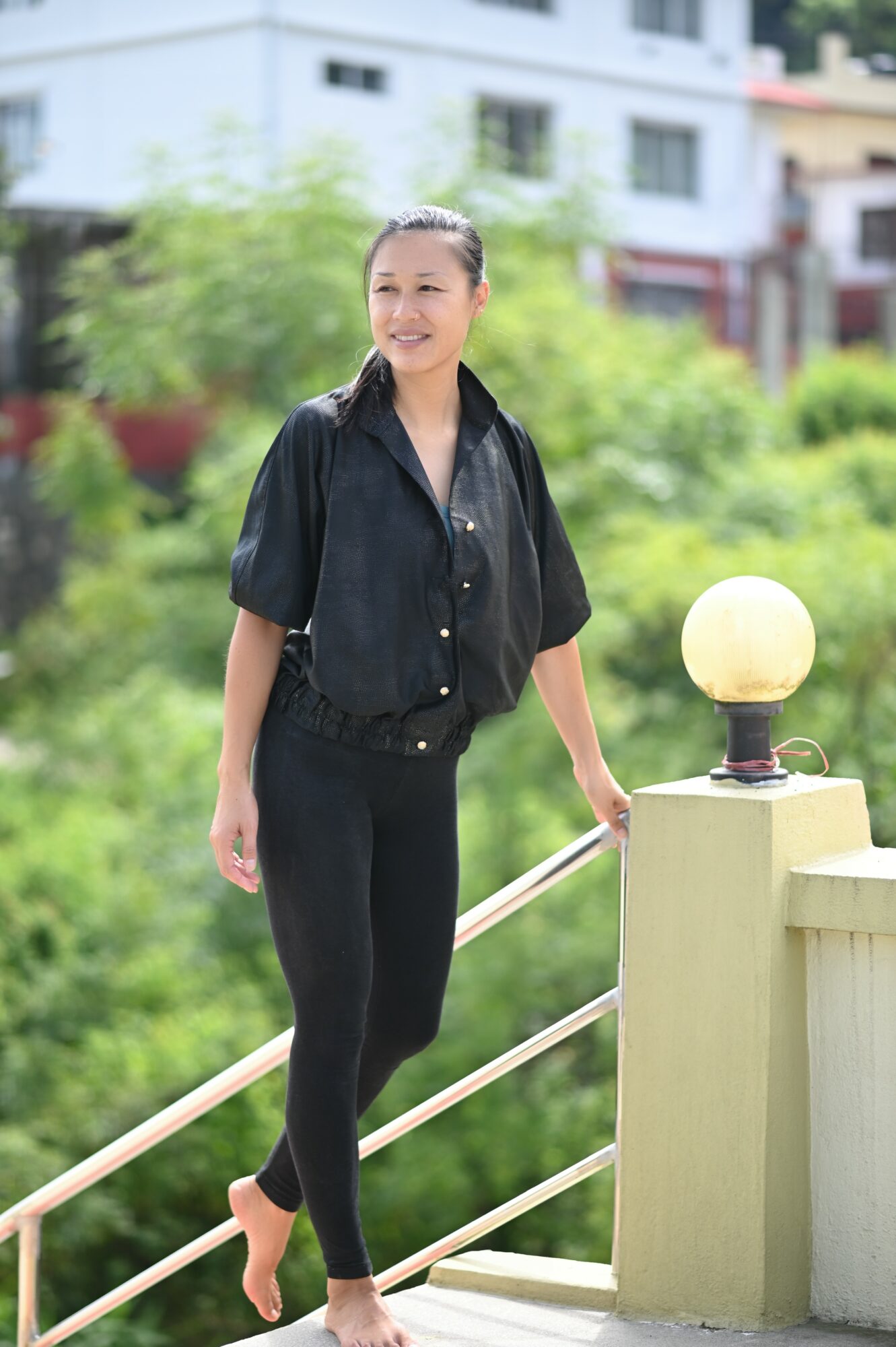 Young woman standing outdoors on a bridge, smiling, wearing black shirt and pants, with greenery and buildings in background.