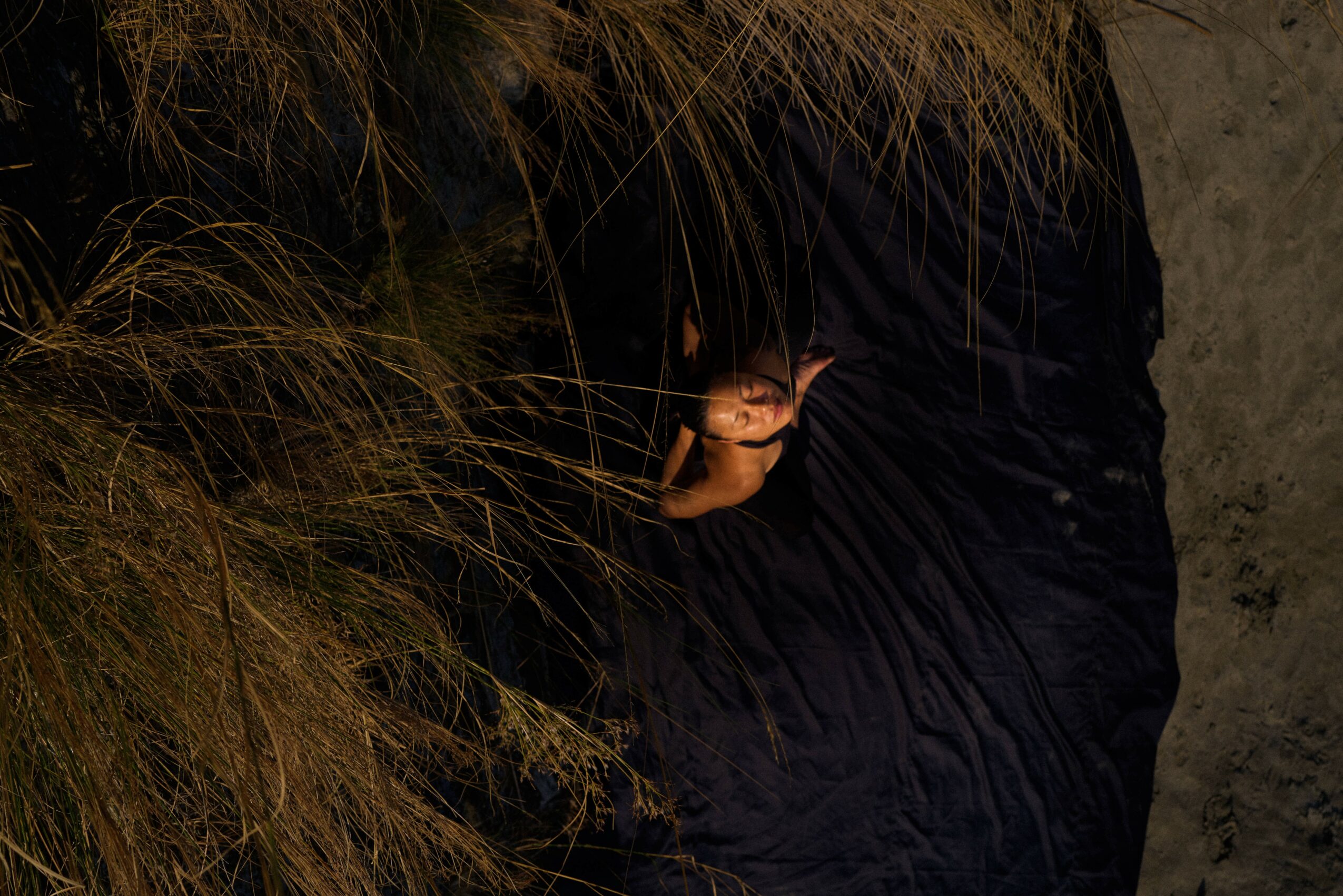 Person with dark clothing holding a small object near tall grass, standing on a dirt surface.