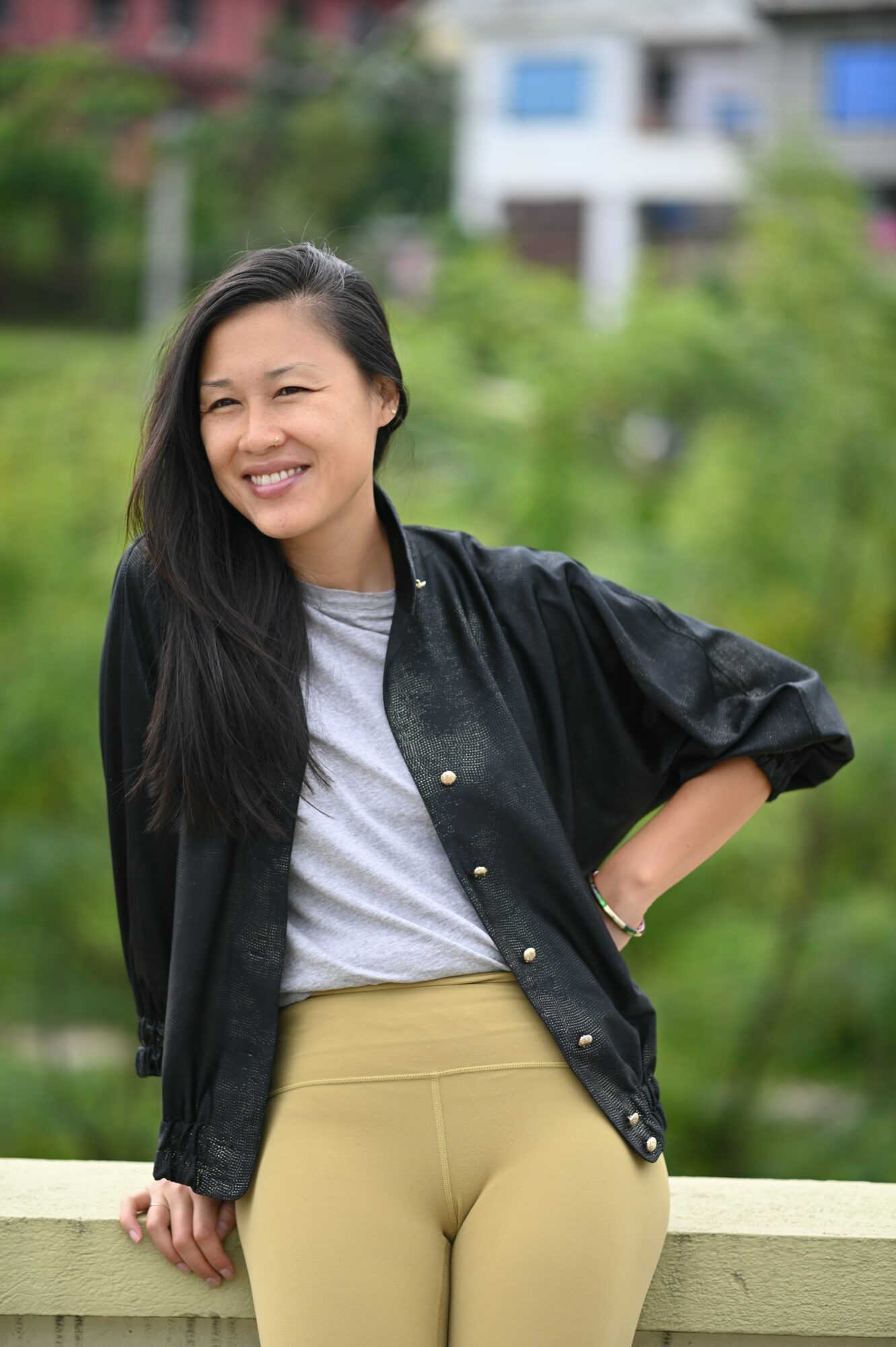 Young woman smiling outdoors, wearing a black jacket and beige pants, with a blurred green background and buildings.