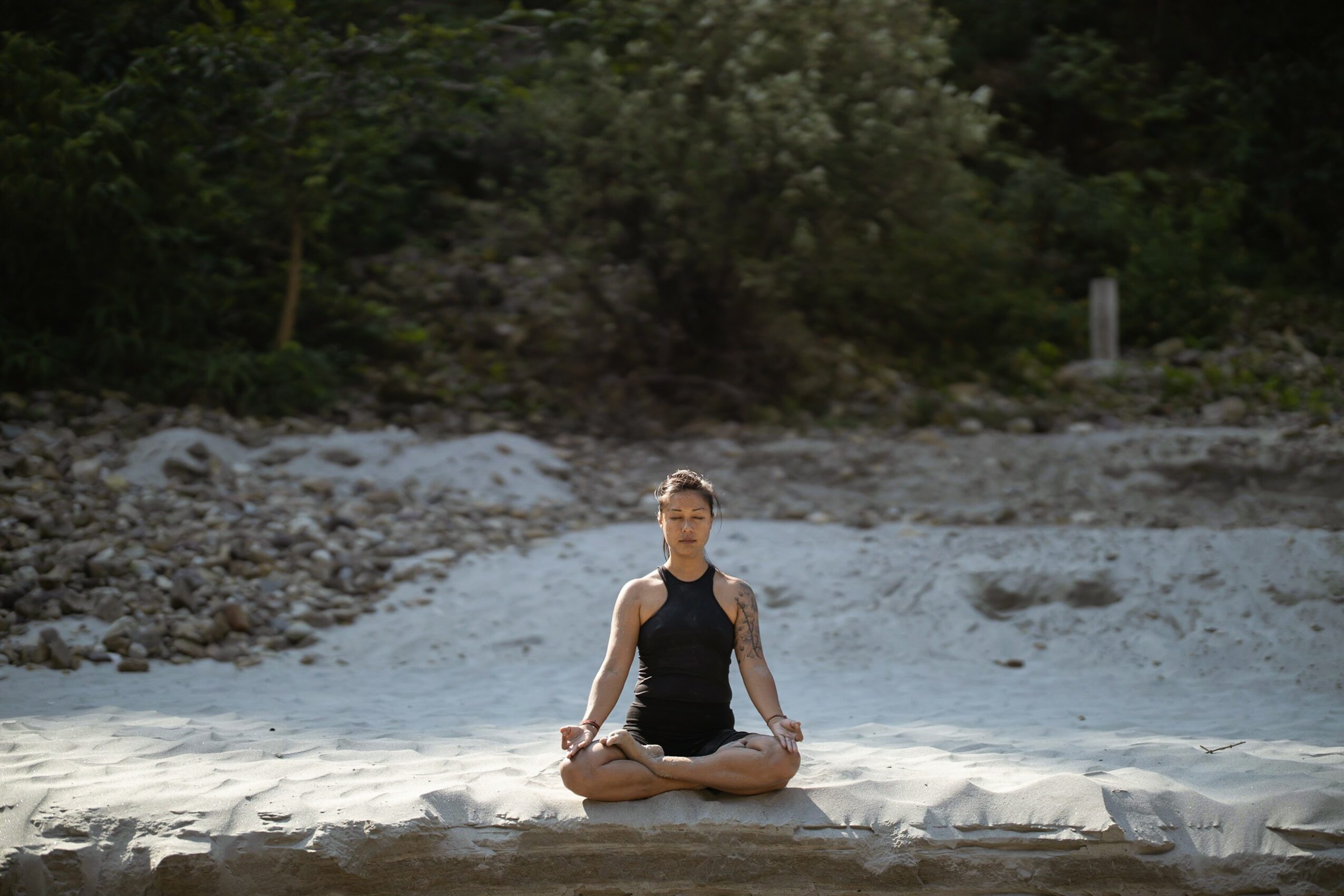 Person sitting cross-legged on a sandy surface outdoors, surrounded by trees and greenery, practicing meditation or yoga.